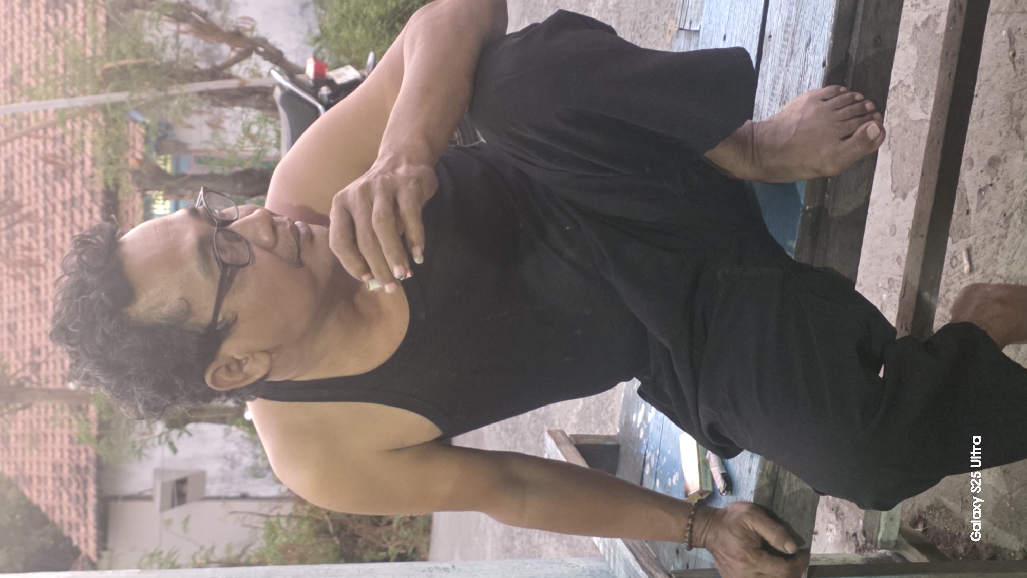 A man is seated outdoors on a blue wooden bench in Juwana, Indonesia. He is wearing a black tank top and black pants. The man is holding a cigarette to his mouth and looking to his right. His hair is curly and dark, and he is wearing glasses. To his left on the bench is a cigarette pack. Behind him, a motorcycle is parked, and a building with a red-tiled roof is visible. The ground is covered in concrete and dirt. The image includes a watermark in the bottom left corner that reads "Galaxy S25 Ultra".