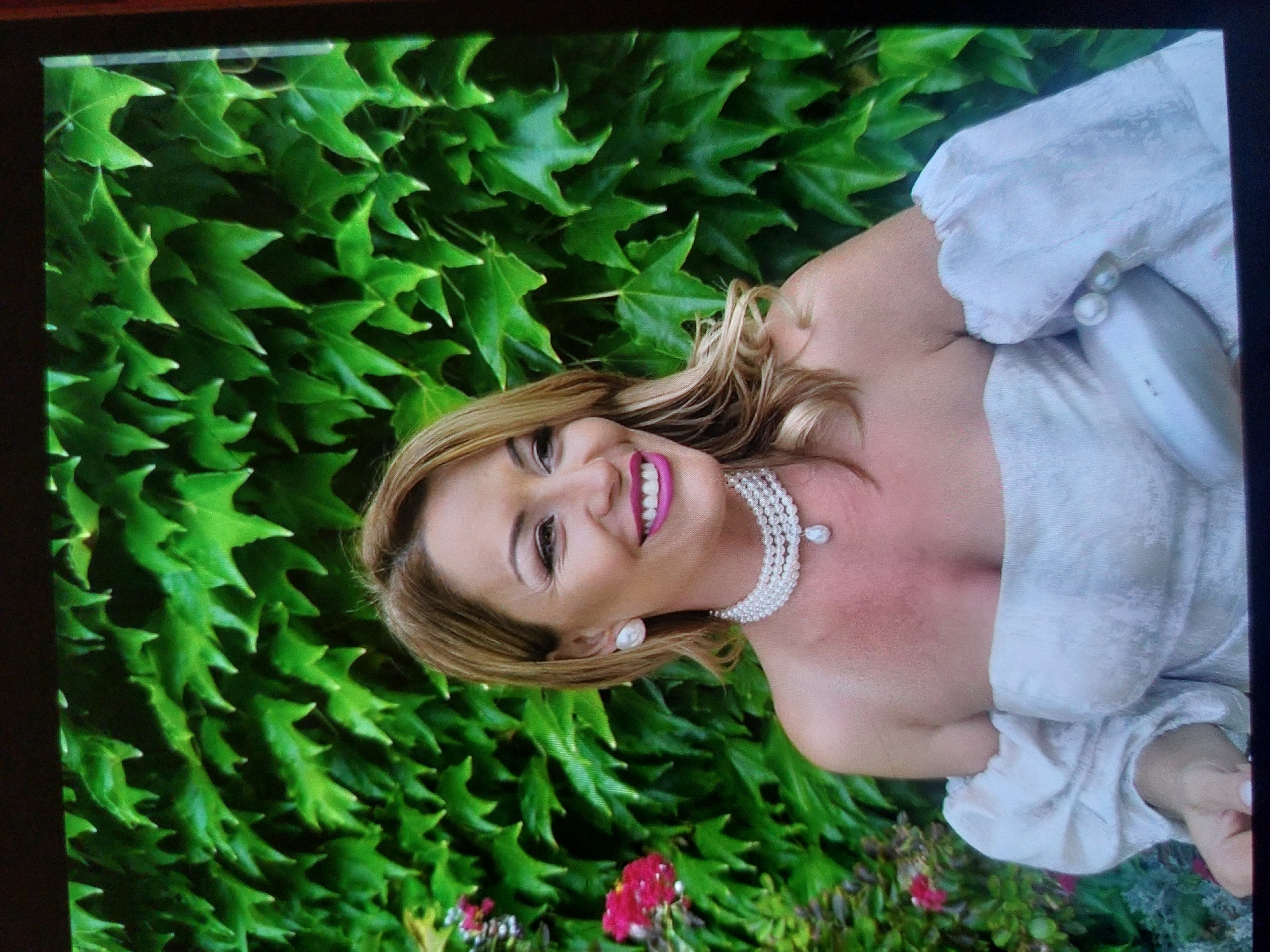 The image displays a cheerful woman posing for a portrait in an outdoor setting, likely in Tsarevo, Bulgaria. She has light-colored hair, possibly blonde or light brown, styled with soft waves, and a broad, joyful smile enhanced by bright pink lipstick. She is adorned with elegant jewelry, including a prominent multi-strand pearl necklace featuring a teardrop pearl pendant, and large pearl stud earrings. Her attire is a light-colored, possibly white or pale grey, strapless or off-the-shoulder garment with gathered fabric details on the upper arms, suggesting a formal or semi-formal dress.
The background is dominated by a dense wall of vibrant green ivy leaves, creating a lush, natural backdrop. Towards the bottom of the frame, glimpses of bright red flowers appear amidst green foliage, indicating a garden or natural environment. The scene is brightly and evenly lit, suggesting it is daytime. The overall mood conveyed is one of happiness and elegance. No text is visible within the image content. The photograph itself appears to be viewed on a digital screen, with a dark frame visible around the edges.