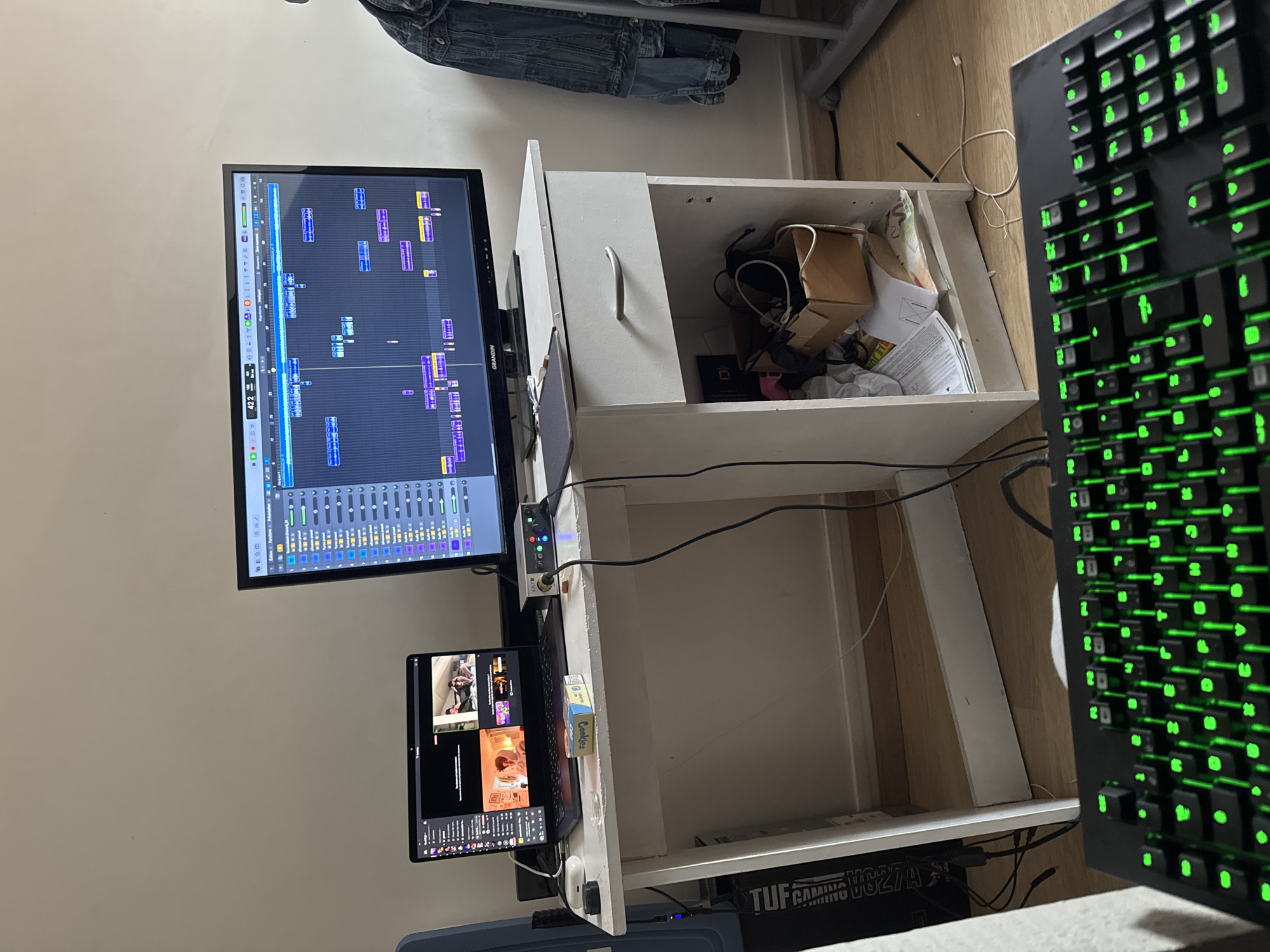 The image shows a workspace with a desk made of white drawers. On the desk, there's a large monitor displaying an audio editing software interface, a smaller screen showing a video, and a black gaming keyboard with green backlighting. The top drawer of the desk is open, revealing a mix of items inside, including a cardboard box, wires, and papers. A box of cookies is positioned near the bottom screen. The bottom shelf of the desk holds a "TUF GAMING VG27A" branded box. The background includes a wall and what appears to be a denim jacket hanging on a rack. There are no people visible in the image. The scene appears to be indoors in a home or office setting. There's no information available that suggests this image was taken in Paris, France.