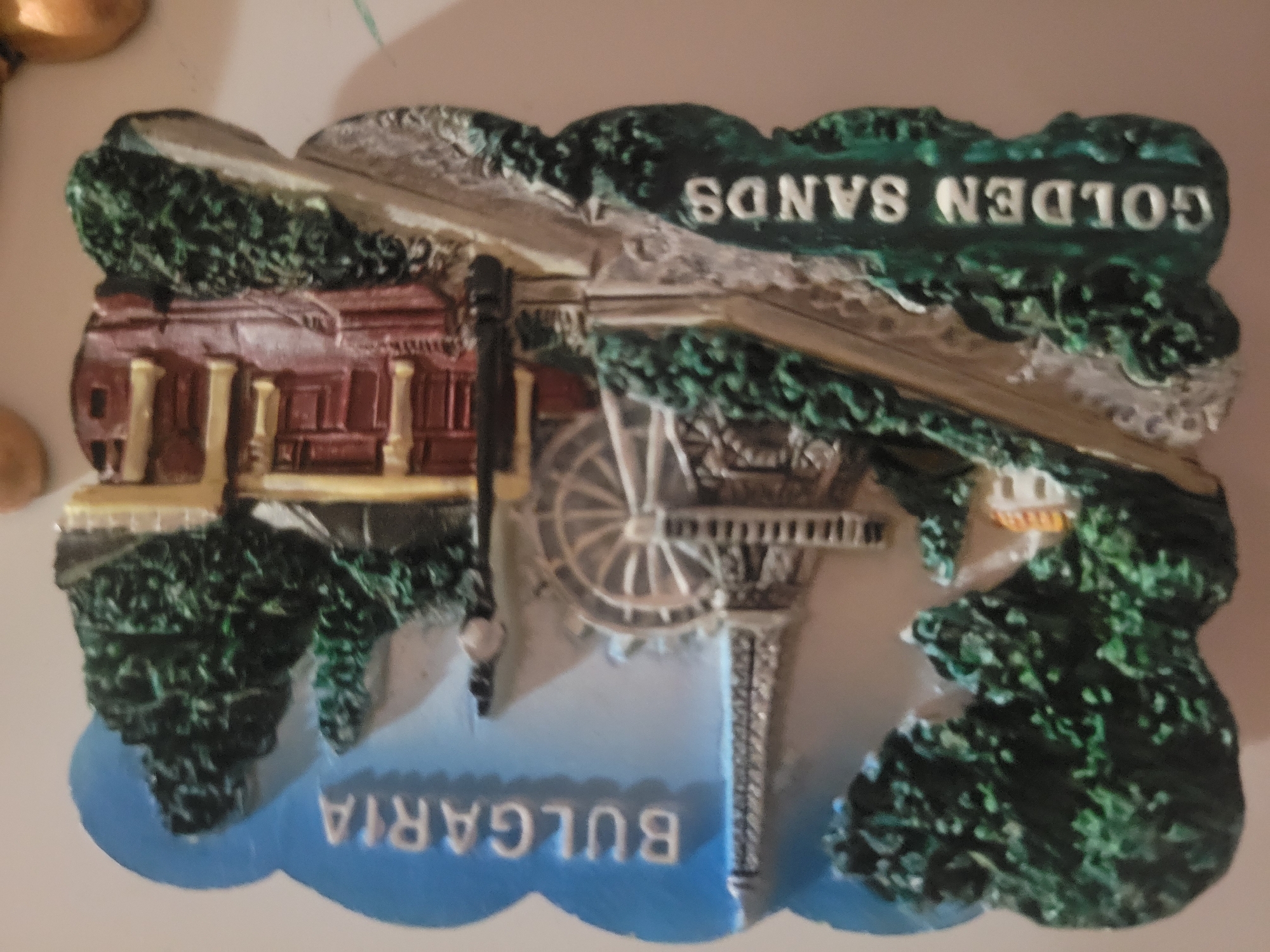The image is a close-up, slightly angled view of a souvenir magnet. The magnet is shaped like a landscape scene with a blue sky at the top and a white surface at the bottom. In the upper part of the magnet, the word "BULGARIA" is written in white, block letters against the blue sky. Below the sky, a miniature Eiffel Tower is depicted, appearing silver and intricately detailed. To the right of the Eiffel Tower, a Ferris wheel is visible. Further to the right, a building with red walls and yellow columns is shown, along with some dark green trees. At the bottom of the magnet, the words "GOLDEN SANDS" are inscribed in white letters on a green, textured surface. The magnet is a three-dimensional representation of a tourist attraction. There are no people or animals present in the image. The scene is a static display and does not depict any ongoing activity.