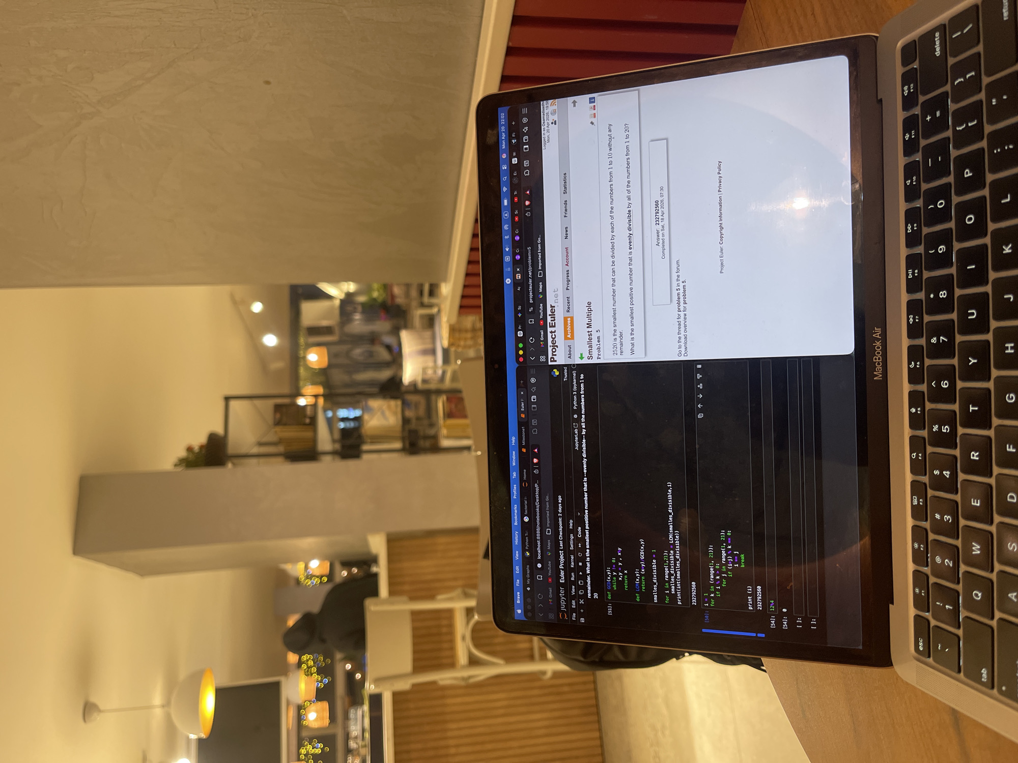 The image captures a MacBook Air laptop open on a light-colored table, likely in an indoor setting such as a café or co-working space in Karabük, Turkey. The background shows warm, artificial lighting from various fixtures, tables, and chairs, with a blurry figure seated in the distance.
The laptop screen displays two main applications. On the left, a Jupyter Notebook environment shows Python code. The code defines functions for Greatest Common Divisor (GCD) and Least Common Multiple (LCM), then uses them to find the "smallest_divisible" number for integers in the range of 1 to 20 (inclusive, using `range(1, 21)`). The output of the Python script is `232792560`. The Jupyter Notebook also indicates "Last Checkpoint: 2 days ago" and "Python 3 (ipykernel)".
On the right side of the screen, a web browser is open to Project Euler Problem 5, titled "Smallest Multiple." The problem asks: "What is the smallest positive number that is evenly divisible by all of the numbers from 1 to 20?" The website displays the correct answer as `232792560` and notes it was "Completed on Sat, 18 Apr 2026, 07:30" by user "Osamadaadhi." The laptop's system clock in the top menu bar reads "Mon Apr 20 22:02". The overall activity appears to be programming and problem-solving, specifically for a mathematical challenge.