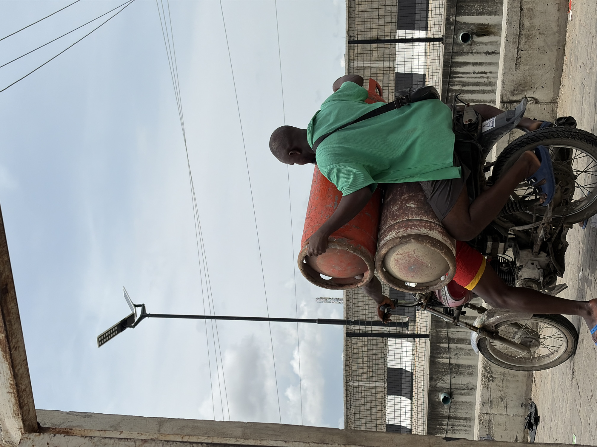 In Igboefon, Nigeria, two men are riding a motorcycle loaded with two large red gas cylinders. The man in the front is driving the motorcycle, wearing a green shirt and dark shorts, with a black bag slung over his shoulder. The man behind him is holding the cylinders, wearing a red and yellow band around his shorts. The gas cylinders are stacked vertically and secured to the back of the motorcycle. The motorcycle is on a paved road, with a concrete barrier and a chain-link fence in the background. A solar-powered street light is visible in the sky, along with power lines and clouds. The scene appears to be an outdoor, possibly industrial or commercial area.