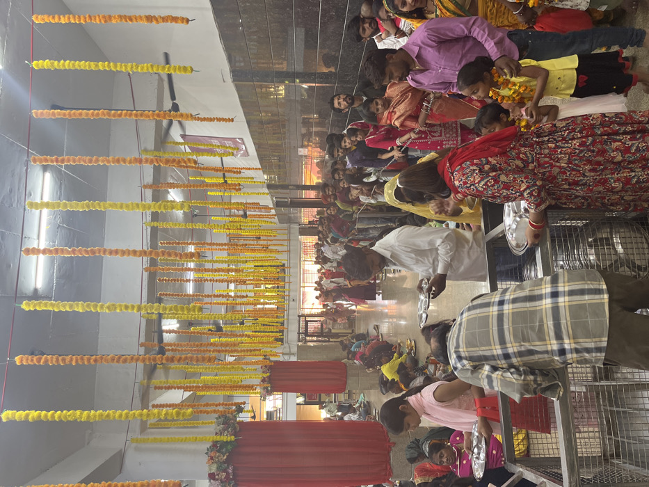 The image shows a crowd of people standing in line at a food distribution event inside a building located at the coordinates 30.723, 76.862, which is in Panchkula, India. The ceiling is decorated with hanging strings of yellow and orange flowers. People are lined up to receive food, while a man at the head of the line is holding a plate, and others behind him are receiving plates. People in line are interacting with each other. The environment is an indoor setting with a tiled floor and a metal cage in the foreground containing empty plates.