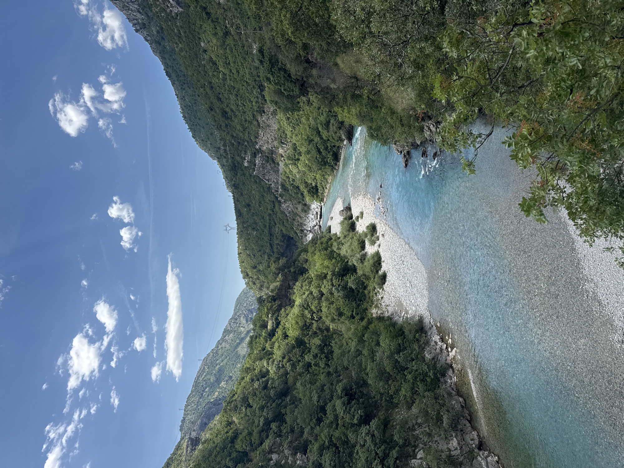 The image depicts a river flowing through a mountainous, forested region in Unknown, Montenegro. The water appears clear and turquoise, with visible pebbles and rocks beneath the surface. The riverbed is wide, consisting of gravelly banks. Lush green vegetation covers the steep slopes of the mountains on either side of the river. The sky is blue with scattered white clouds. Power lines are visible crossing the valley.