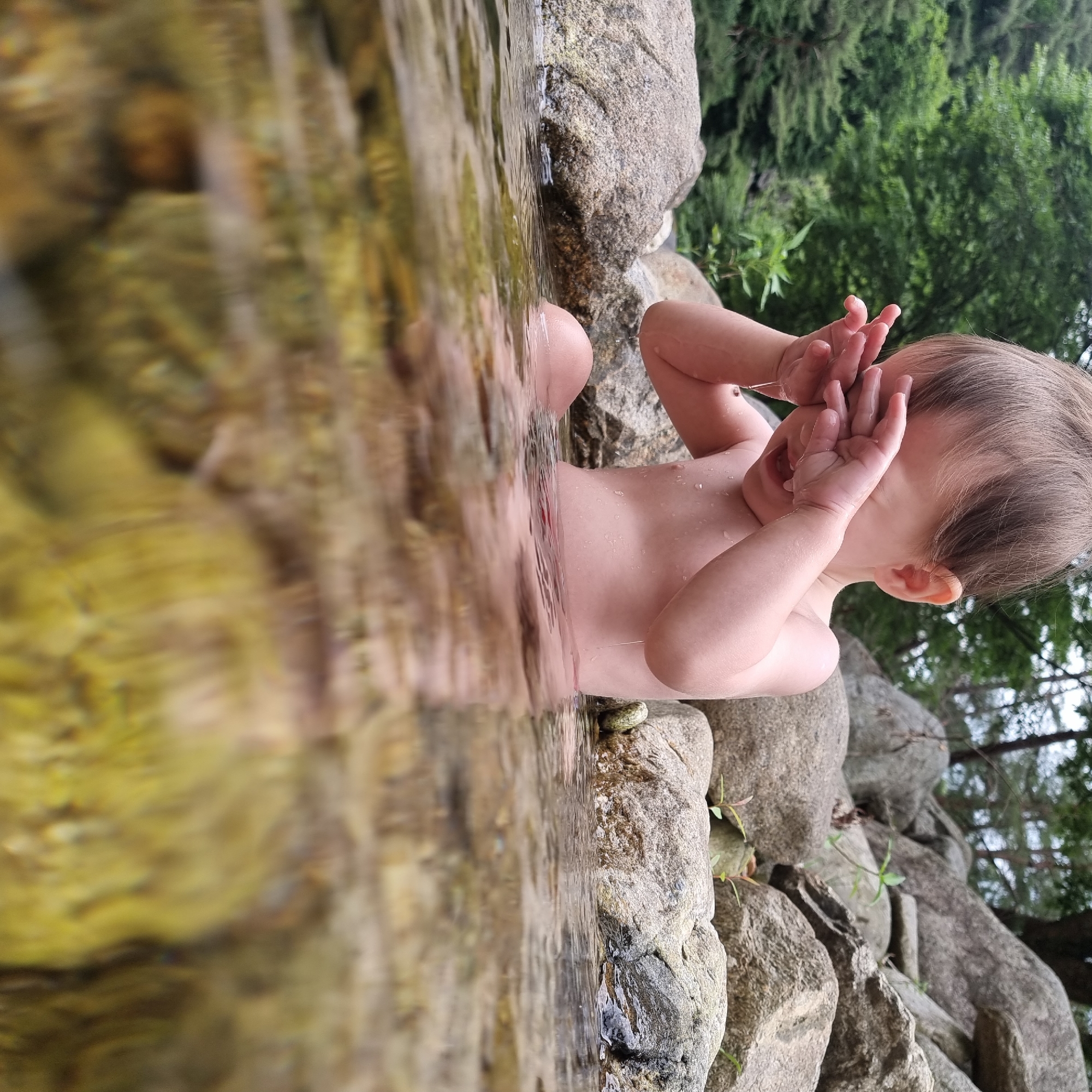 A baby sits in shallow, clear water near the bank of a stream. The baby's hands are held up to their face, covering their eyes, and water drips from their fingers. The baby is partially submerged, with their torso and legs visible above the water. The surrounding environment consists of large, moss-covered rocks and lush green trees. The water is shallow enough to see the rocky bottom with some greenish algae.
