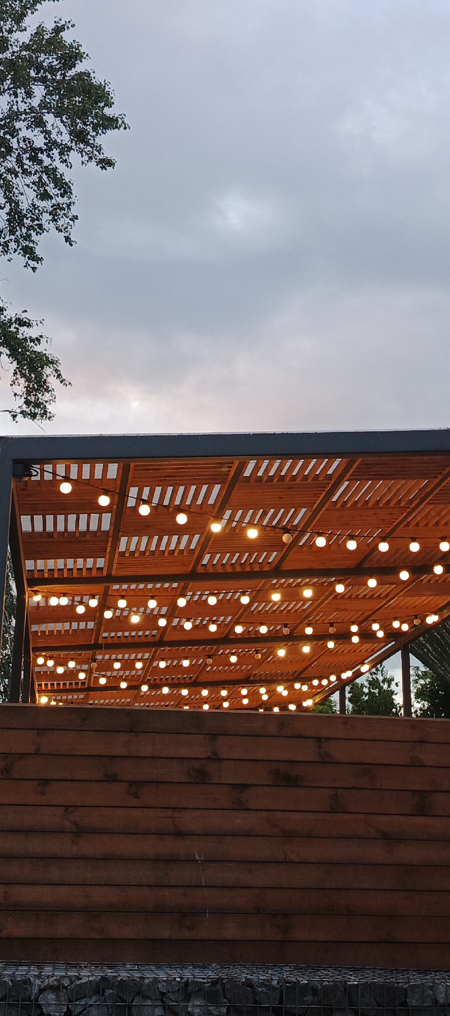 The image shows an outdoor seating area under a wooden pergola. The pergola has a slatted wooden roof, and strings of round, illuminated light bulbs are suspended beneath it, casting a warm glow. The background consists of a cloudy, overcast sky and some greenery, including tree branches in the upper left corner. In the foreground, there is a horizontal wooden fence, and below that, a gabion wall filled with stones. The scene appears to be set in a public space, possibly a cafe or restaurant. The image does not contain any people or depict any specific actions. The location is stated to be Votkinsk, Russia.