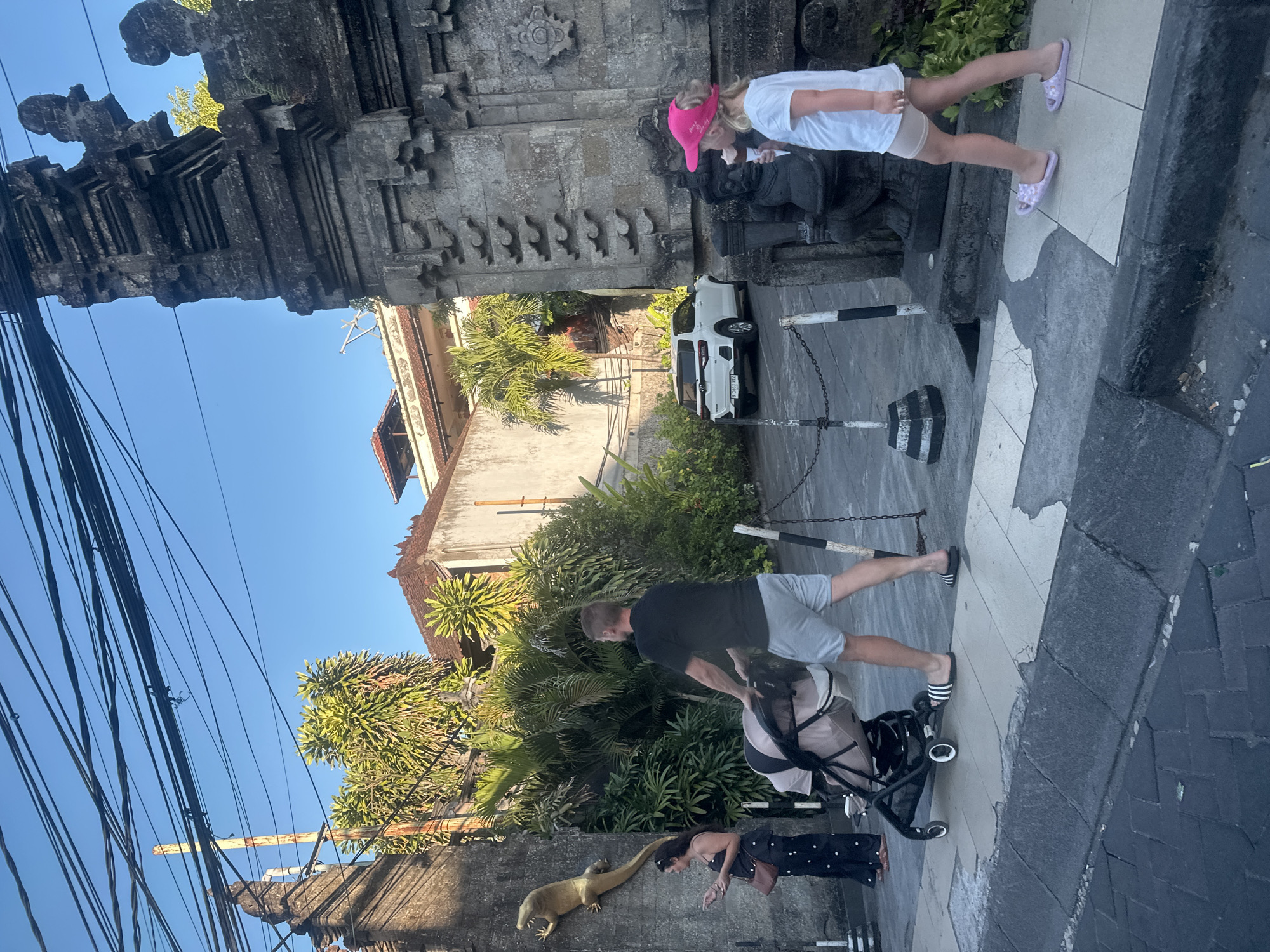 In Kuta, Indonesia, a man pushes a stroller on a sidewalk in front of a stone archway. The man is wearing a black t-shirt and grey shorts. The stroller is beige and black. To the left of the man, a woman in a black and white polka dot dress stands near a golden lizard statue mounted on a stone wall. Behind them, lush green foliage and buildings with tiled roofs are visible. To the right of the archway, a young girl wearing a pink hat and a white t-shirt walks away from the viewer, holding an ice cream cone. A white SUV is parked in the background. Overhead, a tangle of electrical wires crosses the clear blue sky.