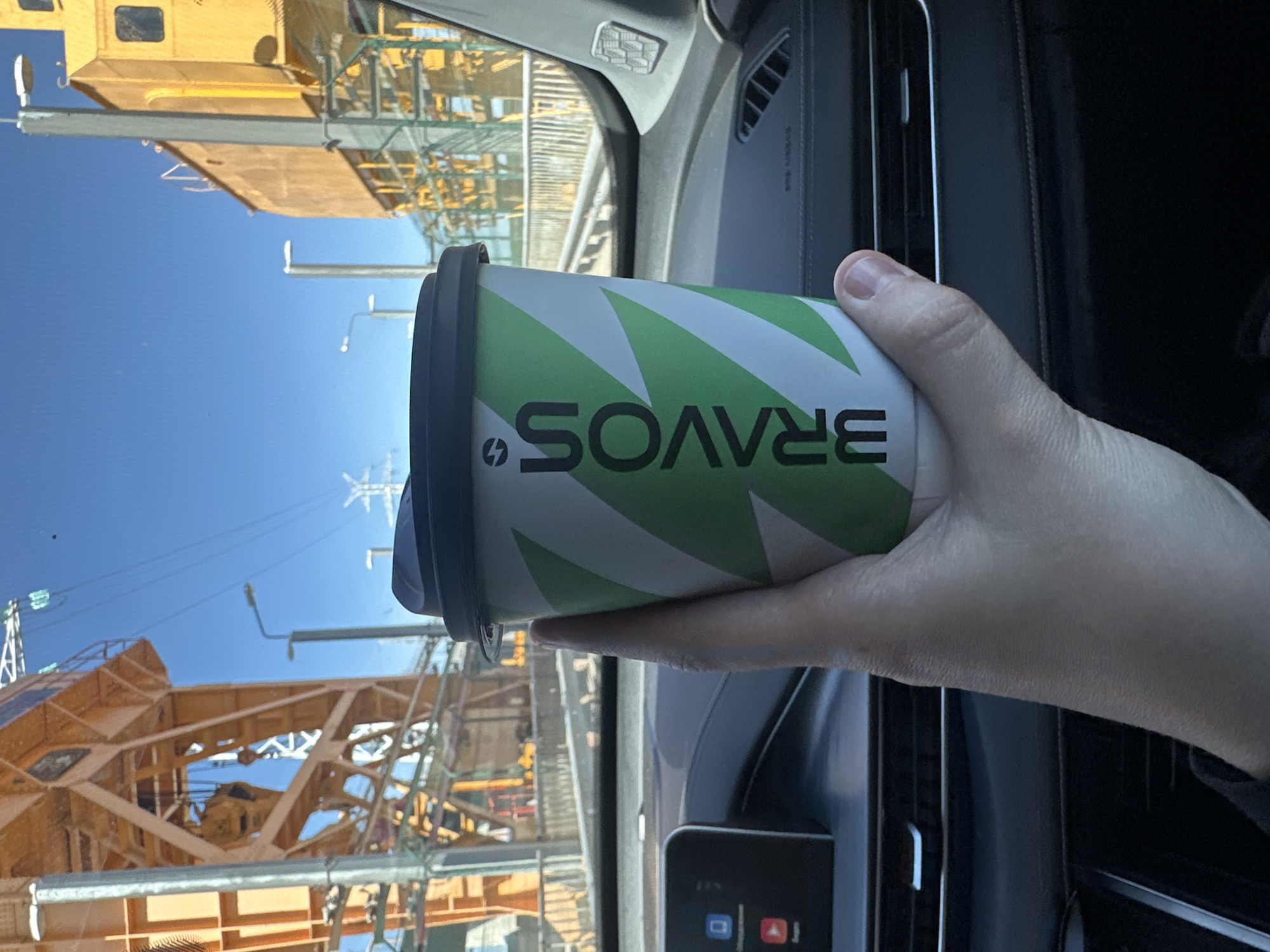 A person inside a vehicle holds a "Bravos" brand paper coffee cup with a dark blue lid. The cup has a green and white chevron pattern. The reflection in the windshield shows what appears to be a construction site with a crane. The dashboard of the vehicle is visible. The location is possibly near a construction site with large cranes, based on what is visible through the windshield. The city is mentioned as Chaikovsky, Russia.