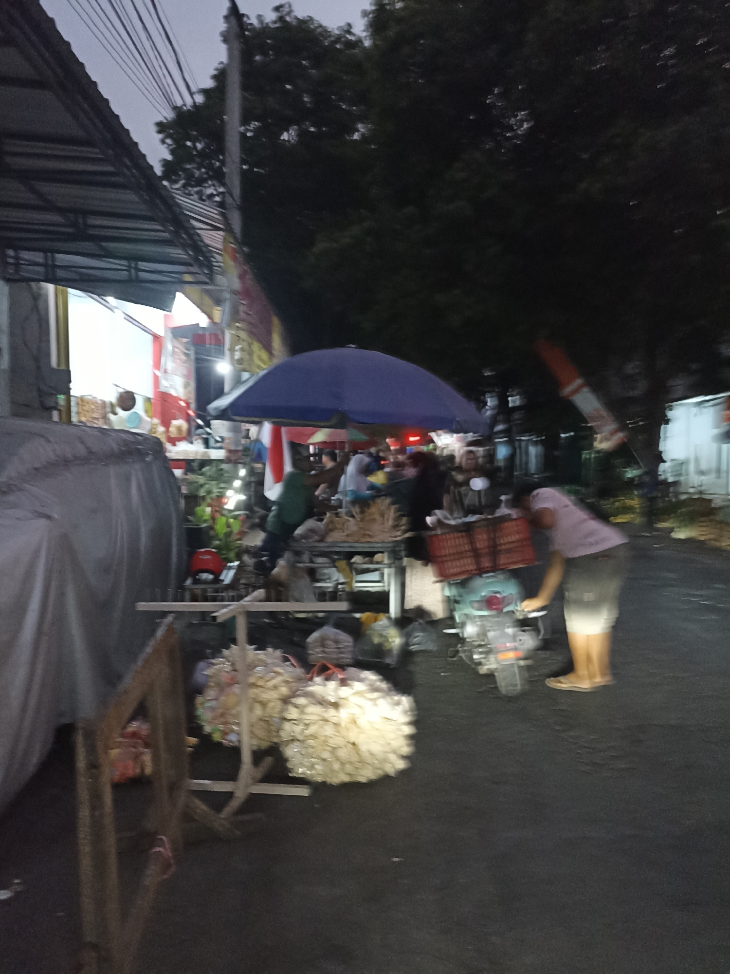 The image depicts a street scene in Ditotrunan, Indonesia, likely in the evening or early morning given the dim lighting and presence of streetlights. In the foreground, a grey tarp covers an object, possibly a vehicle or a stall. To the left, a wooden structure with vertical supports is visible. Beyond this, a street market is set up with various stalls and vendors. One stall features a blue umbrella, under which food items and supplies are displayed on tables. A person in a green shirt is interacting with items at this stall. Further to the right, a light blue scooter is parked, with a person in a pink shirt and grey shorts leaning over its handlebars. The scooter has a red crate on its back filled with goods. In the background, trees and buildings are visible, along with power lines overhead. The ground is asphalt. The overall impression is that of a busy, albeit dimly lit, street market.
