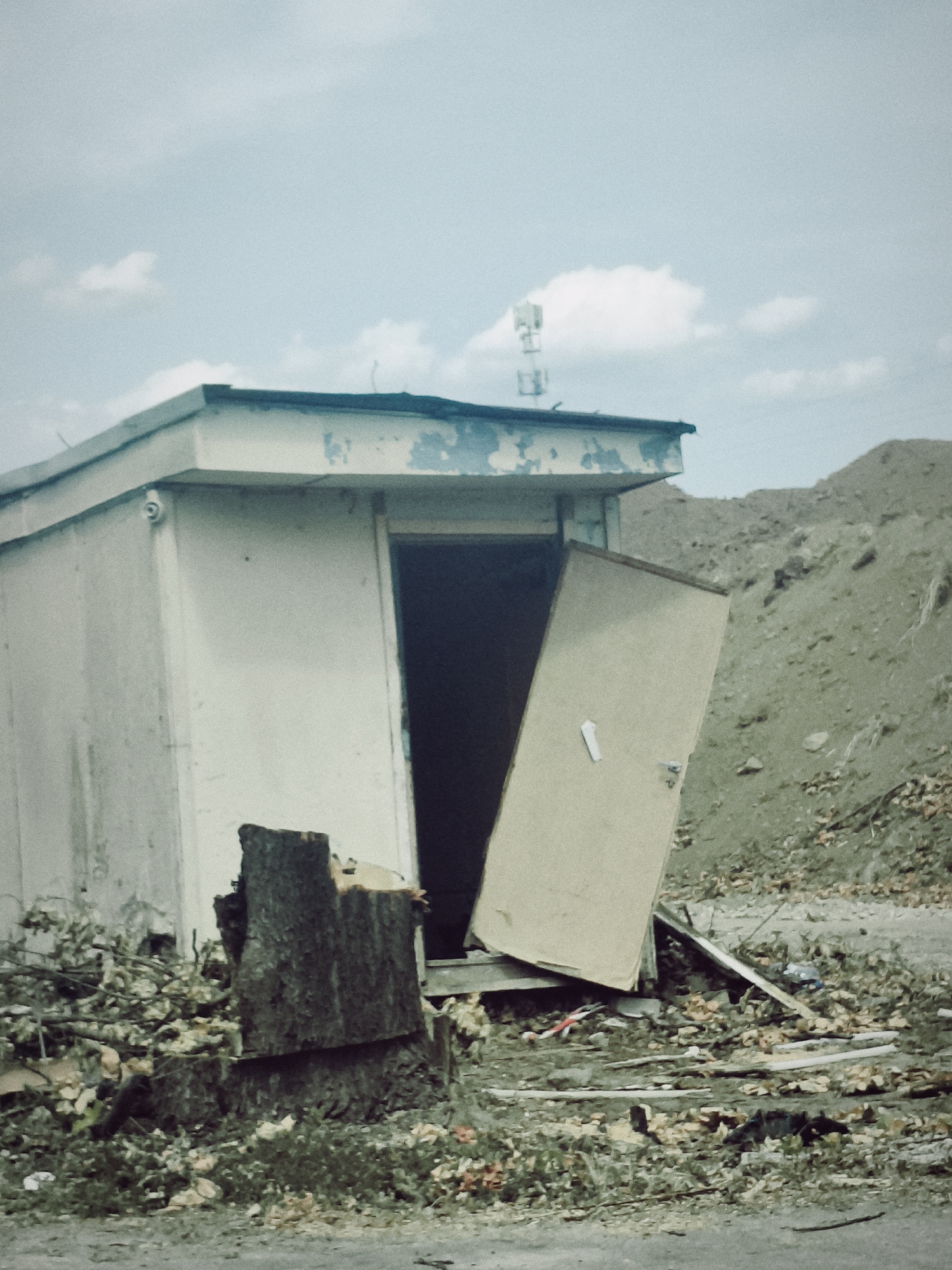 A lone, weathered, white portable building with a slightly damaged, light-colored door ajar is situated in an unkempt, possibly abandoned or construction-adjacent outdoor environment. The building's roof has peeling blue trim. Debris, including branches and what appears to be construction waste, is scattered around the base of the structure. A large, rough-hewn tree stump is positioned in front of the building's left side. In the background, a barren, sloped hill covered in rocks and sparse vegetation dominates the landscape, suggesting a remote or undeveloped area. The sky above is overcast with scattered clouds. A distant cell tower with antennas is visible on the horizon above the building. The scene appears to be located in Palmiry, Poland.