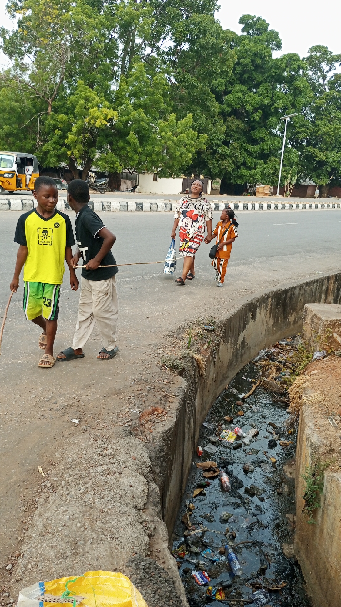In Adankolo, Nigeria, two young boys walk along a sidewalk adjacent to a polluted drainage canal. One boy holds a stick. Behind them, a woman and a young girl walk on the road. A yellow-colored auto rickshaw is visible in the background. The environment includes trees and buildings. The drainage canal contains trash and stagnant water.