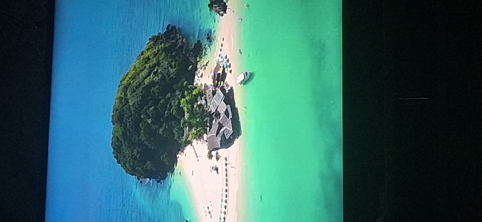 This is an aerial photograph of a tropical island with a white sand beach, lush green trees covering the island's interior, and clear turquoise and blue waters. There are several small buildings with dark roofs situated on the beach, appearing to be part of a resort or restaurant complex. Numerous beach umbrellas and lounge chairs are set up on the sand. Several small boats are visible in the water near the shore, and one larger white boat is anchored further out. The environment is serene and appears to be a popular tourist destination. There is no discernible connection to the city of Bila Tserkva, Ukraine, as the image depicts a tropical island setting.
