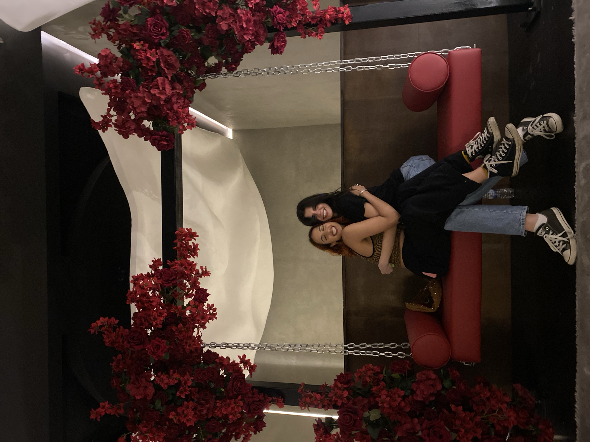 Two women are posed lying on a hanging, cushioned red bench suspended by chains, under a structure decorated with red flowers. One woman has long dark hair and is wearing a black top and blue jeans. The other woman has medium-length red hair, is wearing a patterned top, a dark jacket, and blue jeans. They are smiling and appear to be in a social setting. The background includes interior design elements and is likely in a public space in Dubai, United Arab Emirates.