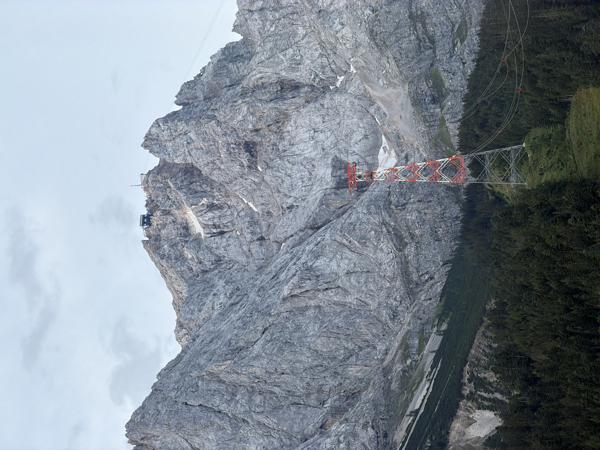 The image depicts a mountainous landscape in Grainau, Germany, likely part of the Zugspitze massif. Dominating the mid-ground is a tall, lattice-style support tower for a cable car system. The tower is painted with a distinctive pattern of red and white sections. Numerous cables stretch diagonally upwards from the tower towards the mountain peak. The mountain itself is characterized by its rugged, grey, rocky terrain, with sparse patches of snow visible near the summit. The lower slopes are covered in a dense coniferous forest. At the very top of the mountain, a building complex, presumably a cable car station or observation point, is visible against a pale, overcast sky. The scene appears to be a daylight shot, with diffuse lighting suggesting cloudy weather.