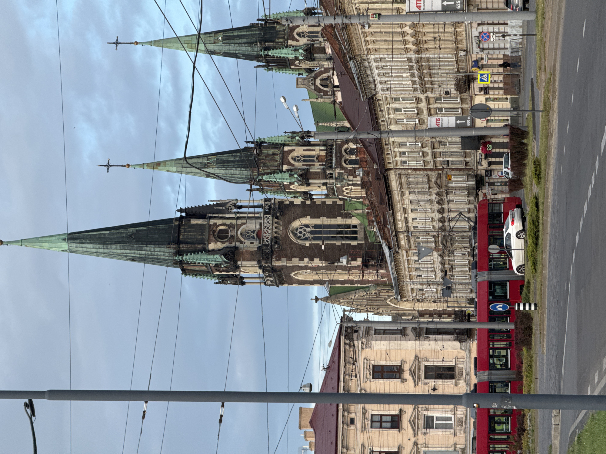 The image shows a street scene in Lviv, Ukraine, dominated by the neo-Gothic spires of the Church of Sts. Olga and Elizabeth. In the foreground, a red tram is stopped at a tram stop, with a white car driving past it. The street is paved with cobblestones, and there are several buildings lining the street, some of which appear to be old and historic. Overhead, a network of electrical wires crisscrosses the sky, supported by utility poles. The sky is overcast, with a few clouds visible. There are a few pedestrians on the sidewalk, and some advertisements are visible on the buildings. The overall impression is of a busy urban environment with a rich architectural heritage.