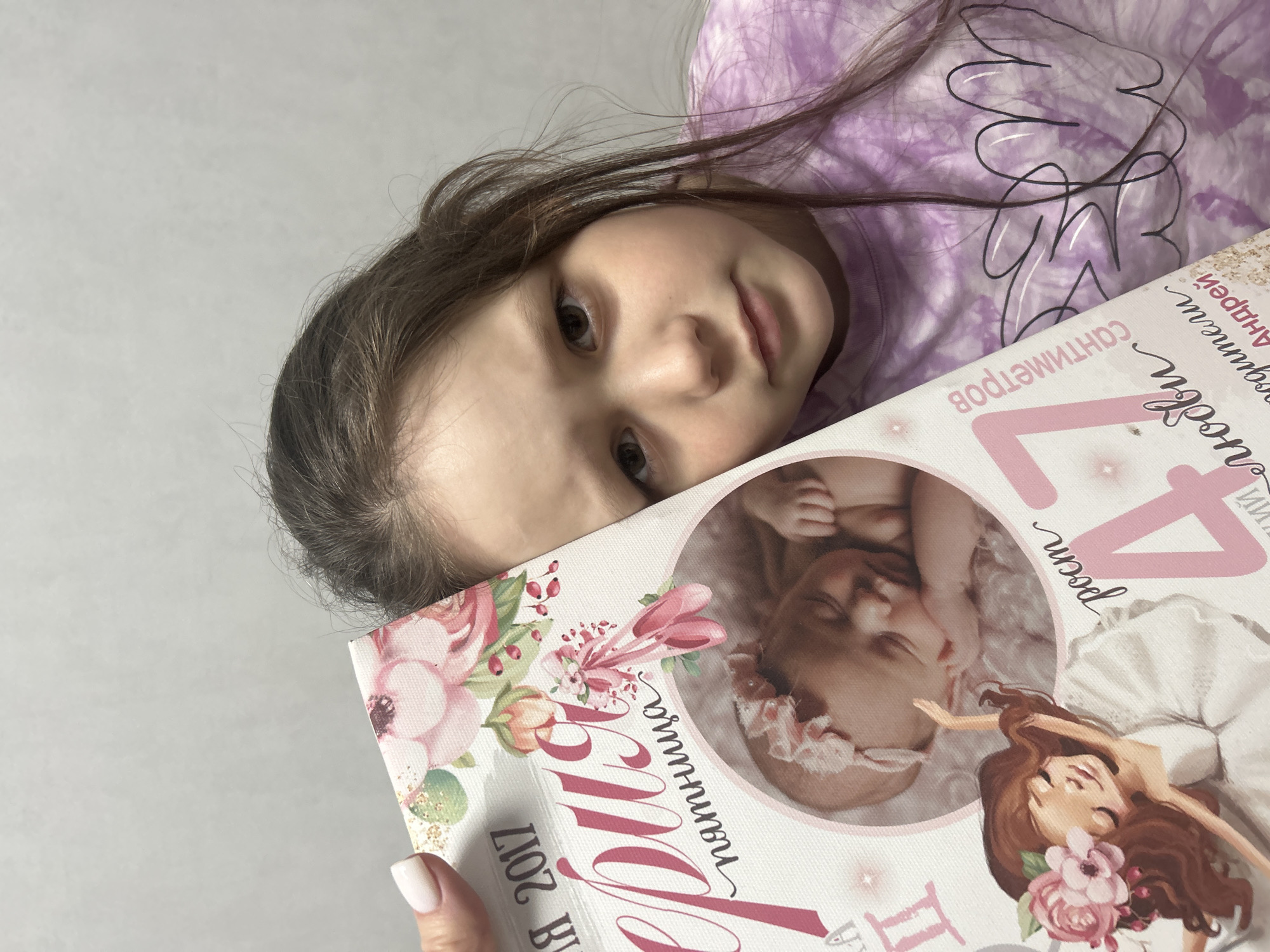 A young girl is holding a book with a baby and cartoon image on the cover. The book also says “Сантиметров Андрей, Родился 4 Росси”. The book mentions an address in Chaikovsky, Russia. The girl is wearing a purple tie-dye shirt and has brown hair. The background is a plain wall.