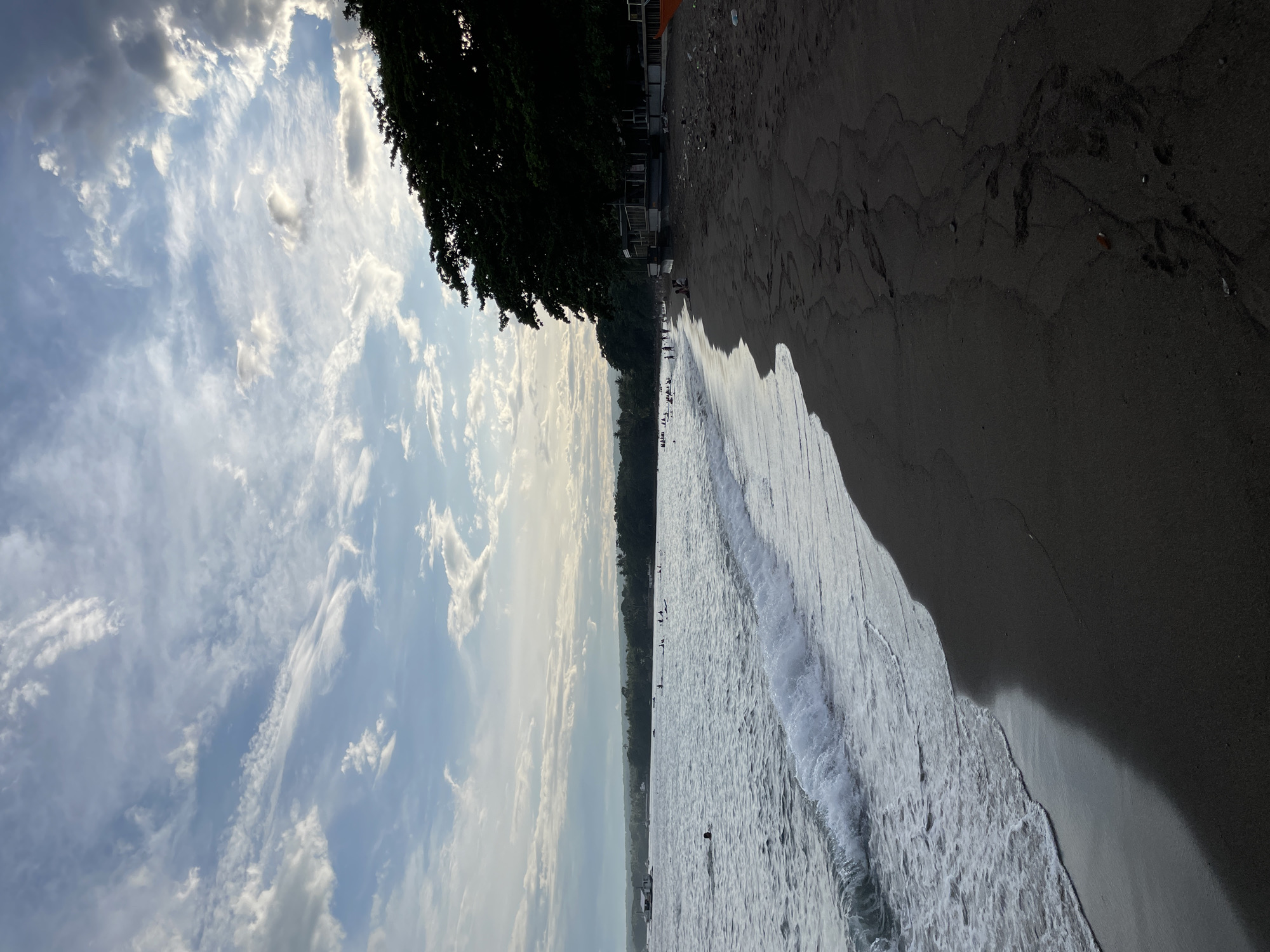 The image depicts a beach scene in Dauin, Philippines, under a sky filled with scattered clouds. The dark sand of the beach occupies the foreground, leading to the ocean. Waves gently break on the shore, with foam spreading across the wet sand. In the water, several people are visible, some swimming and others engaged in water activities. A boat is also present in the distance. Along the edge of the beach, a line of trees provides shade, and structures that appear to be huts or small buildings are situated beneath them. The overall atmosphere is serene, suggesting a leisurely day at the beach.
