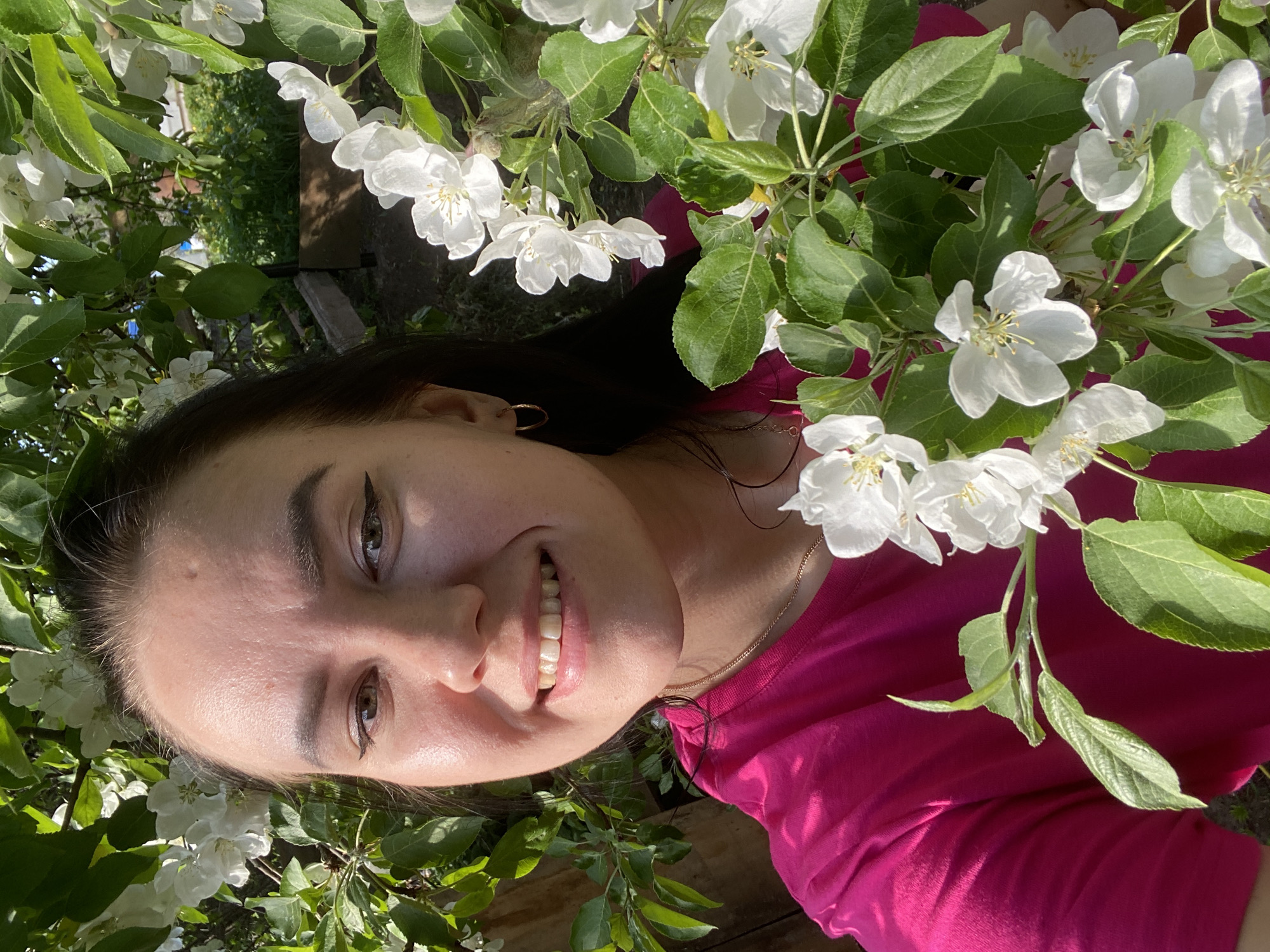 The image depicts a young woman with dark hair and light skin smiling at the camera. She is surrounded by white blossoms and green leaves, suggesting she is outdoors, possibly in an orchard or garden. She is wearing a pink top and a gold necklace is visible. Her eyes are lined with eyeliner and she has a gold hoop earring. The light suggests it is daytime. There is no specific information in the image to indicate that it was taken in Chaikovsky, Russia.