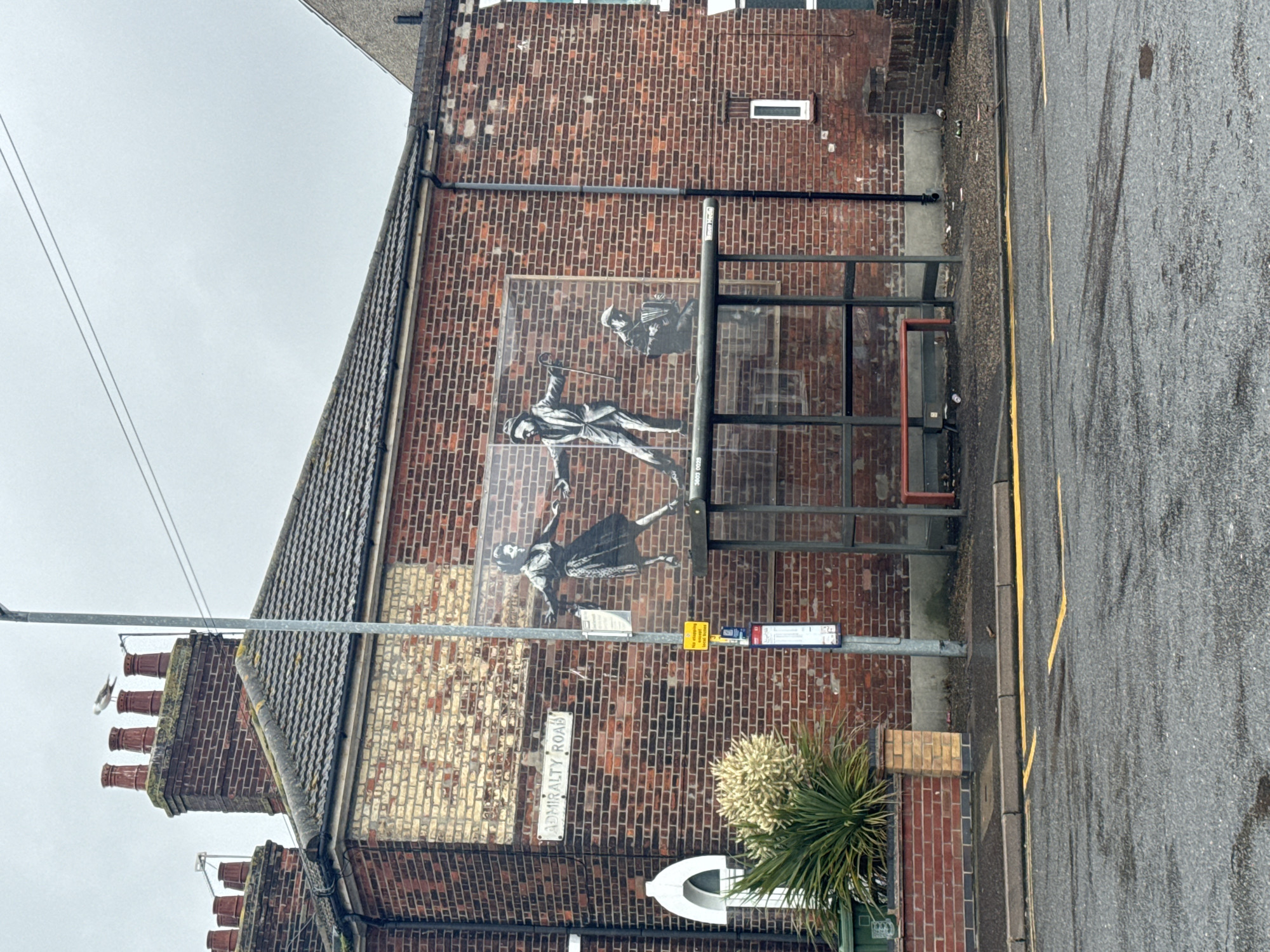 The image captures a street scene in Great Yarmouth, United Kingdom, focusing on a brick building with a large mural depicting two figures dancing and another playing an accordion. The mural is partially obscured by a bus shelter with a red bench. A street sign reading "ADMIRALTY ROAD" is visible on the building. The environment is overcast, with wet asphalt and yellow road markings. Several beer cans are scattered on the ground near the bus shelter. The building's architecture suggests a residential or commercial property, with multiple chimneys on the roof and a distinct arched window. The overall impression is one of urban decay mixed with artistic expression.