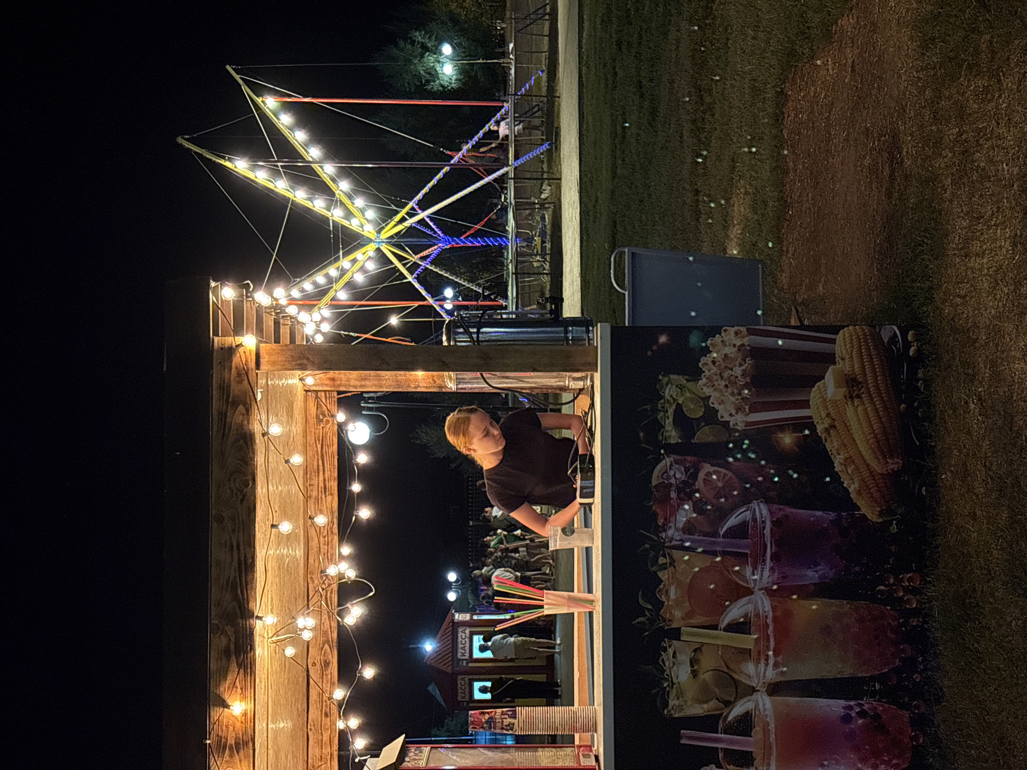 At night, a young woman with blonde hair tied in a ponytail is working behind a concession stand in Sol-Iletsk, Russia. She is wearing a dark t-shirt and is looking down at a device she is holding in her hands, possibly a card reader, as she prepares a drink. The concession stand is decorated with string lights and displays images of colorful drinks, popcorn, and corn on the cob. In the background, people are gathered, and a bungee jumping attraction with illuminated poles and cables is visible. The ground is covered with grass and a dirt path.