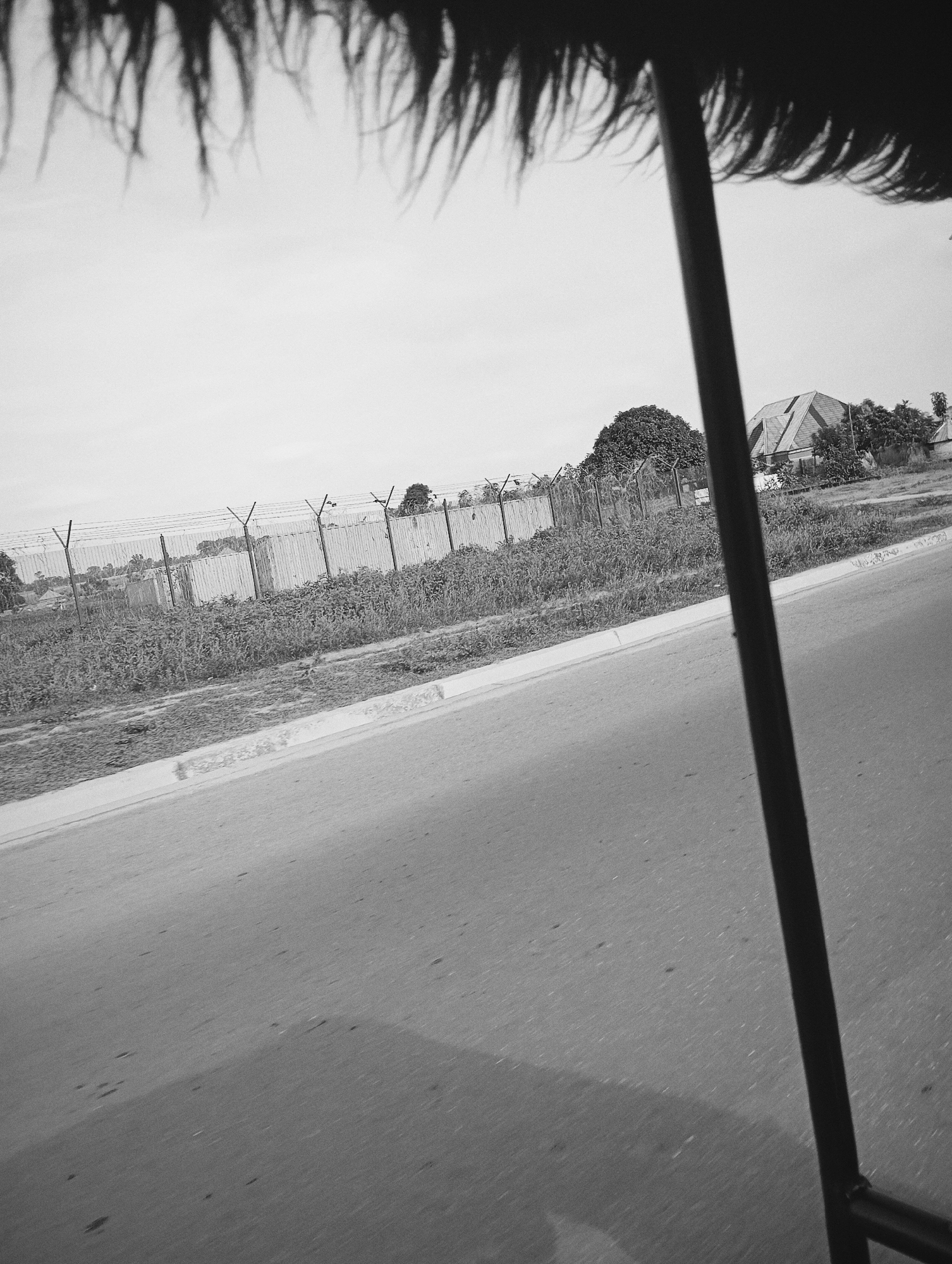 The image, taken from a moving vehicle in Jalingo, Nigeria, shows a roadside scene. In the foreground, a dark, blurry object, possibly a part of the vehicle's structure or canopy, hangs over the top of the frame. A thin, dark vertical pole, likely a support for the canopy, runs along the right side of the image.
The road itself occupies the lower portion of the frame, appearing as a paved surface with a slight incline. The road surface has a grainy texture and shows some scattered small marks. A white concrete curb separates the road from the verge.
Beyond the curb, a strip of dense, overgrown vegetation is visible, consisting of various plants and grasses. Behind the vegetation, a tall fence runs horizontally across the middle ground. This fence appears to be made of metal posts supporting barbed wire, with corrugated metal sheeting in sections.
Further in the background, a cluster of trees is present, and behind them, a building with a distinctive sloped roof is visible. The sky is overcast and pale, dominating the upper portion of the image behind the foreground elements. The overall impression is of a somewhat rural or undeveloped roadside environment.