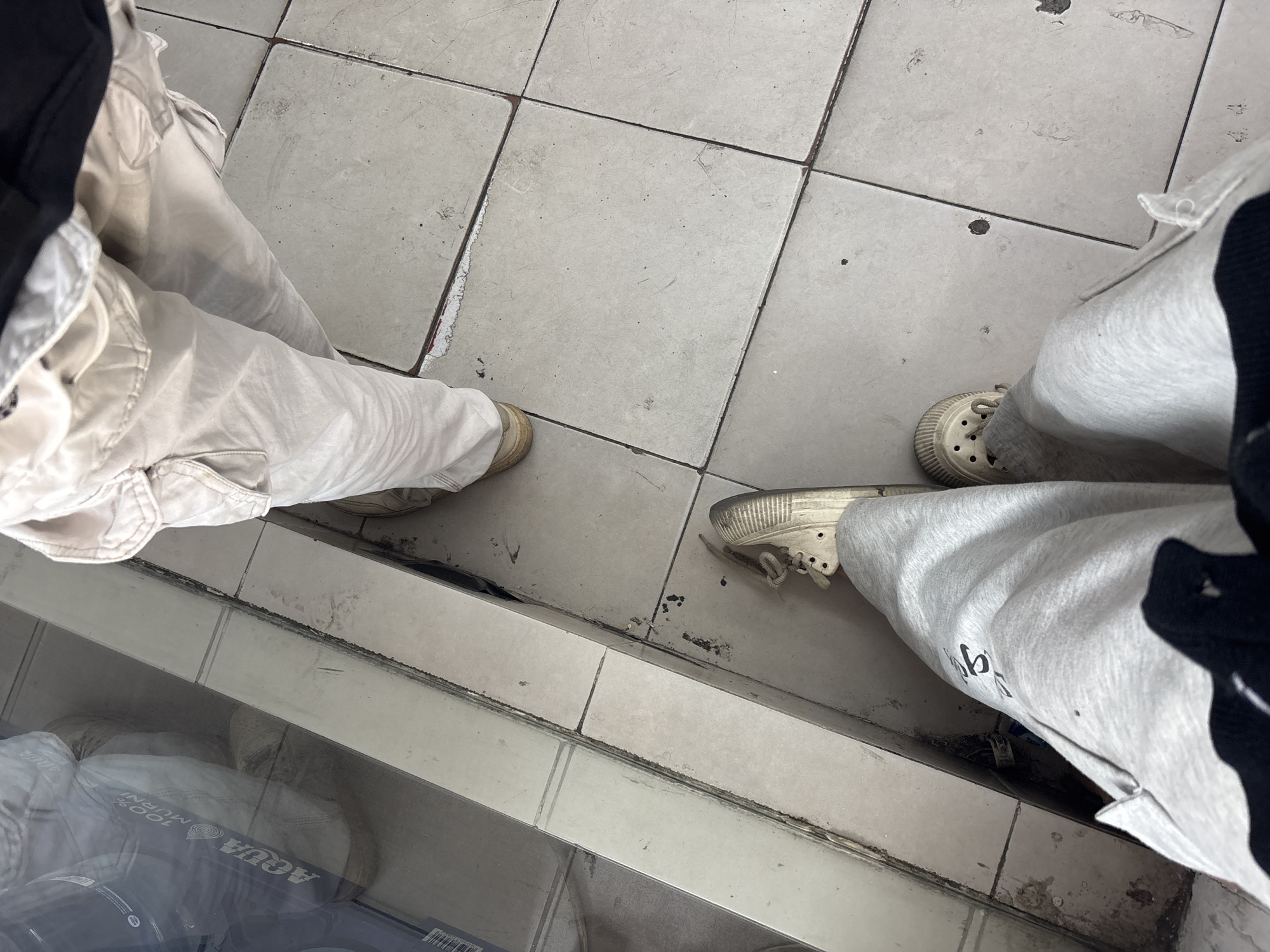 The image shows a first-person perspective of two individuals standing on a tiled floor. The floor is light-colored and appears to be in a somewhat worn condition, with some visible dirt and grime. To the left of the frame, a reflective surface, likely a glass display or window, shows the reflection of one of the individuals, wearing light-colored cargo pants and beige shoes. Text on a bottled drink visible in the reflection reads "AQUA 100% MURNI." The foreground shows the legs of another individual wearing light gray sweatpants and beige clogs. The overall environment suggests an indoor setting, possibly a shop or a public space within North Jakarta, Indonesia, given the presence of the "AQUA" brand, a popular bottled water in Indonesia.