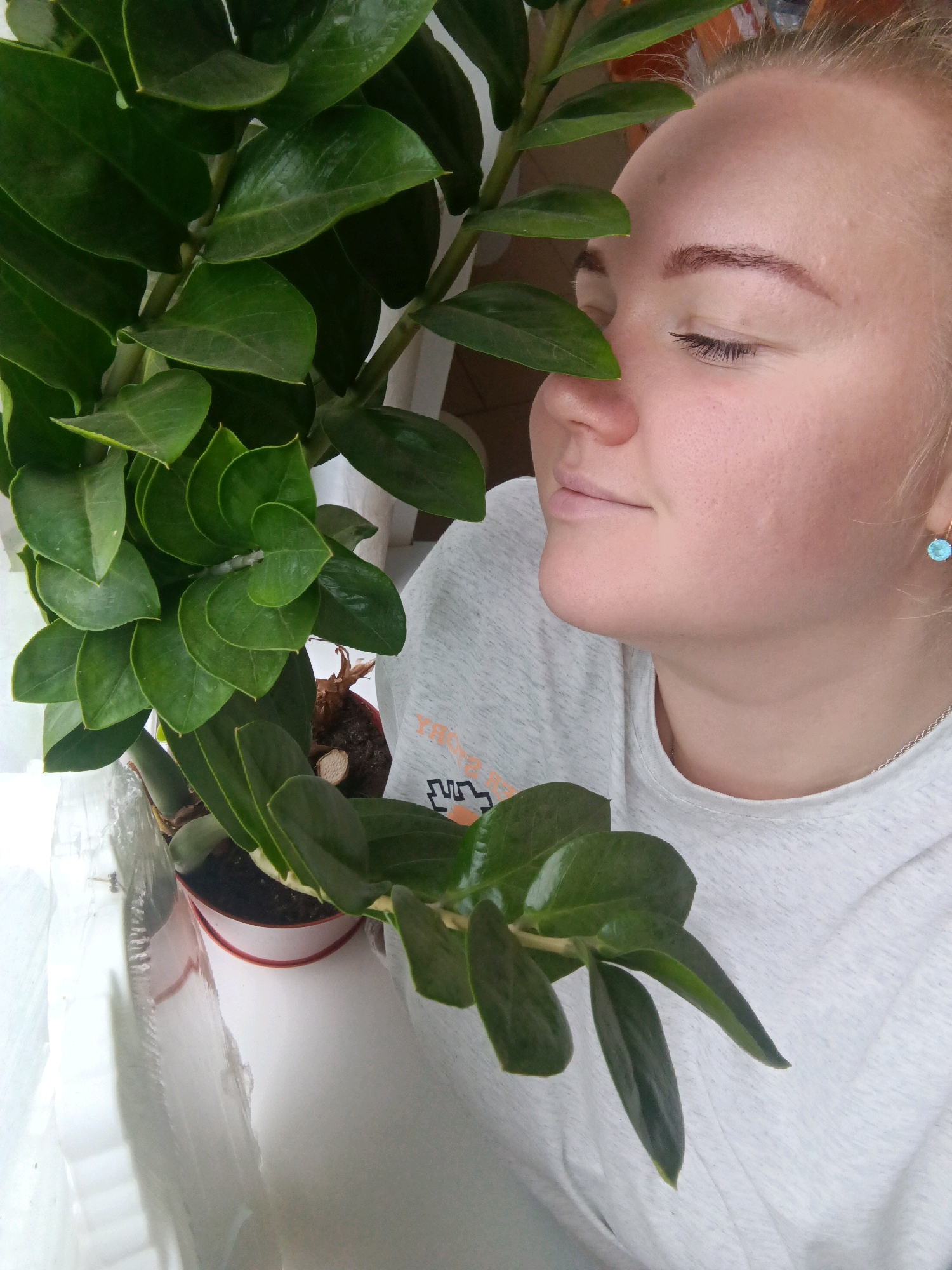 A young woman is shown from the chest up, with her eyes closed and her face turned slightly to the right, smelling a plant with large, dark green leaves. The plant is in a small pot and is positioned in front of her face. She is wearing a light gray t-shirt and a light blue earring in her right ear. The background is softly lit and appears to be an indoor setting. There are no identifiable landmarks or elements that suggest a specific location within the city of Фоки, Russia. The overall mood of the image is peaceful and serene.