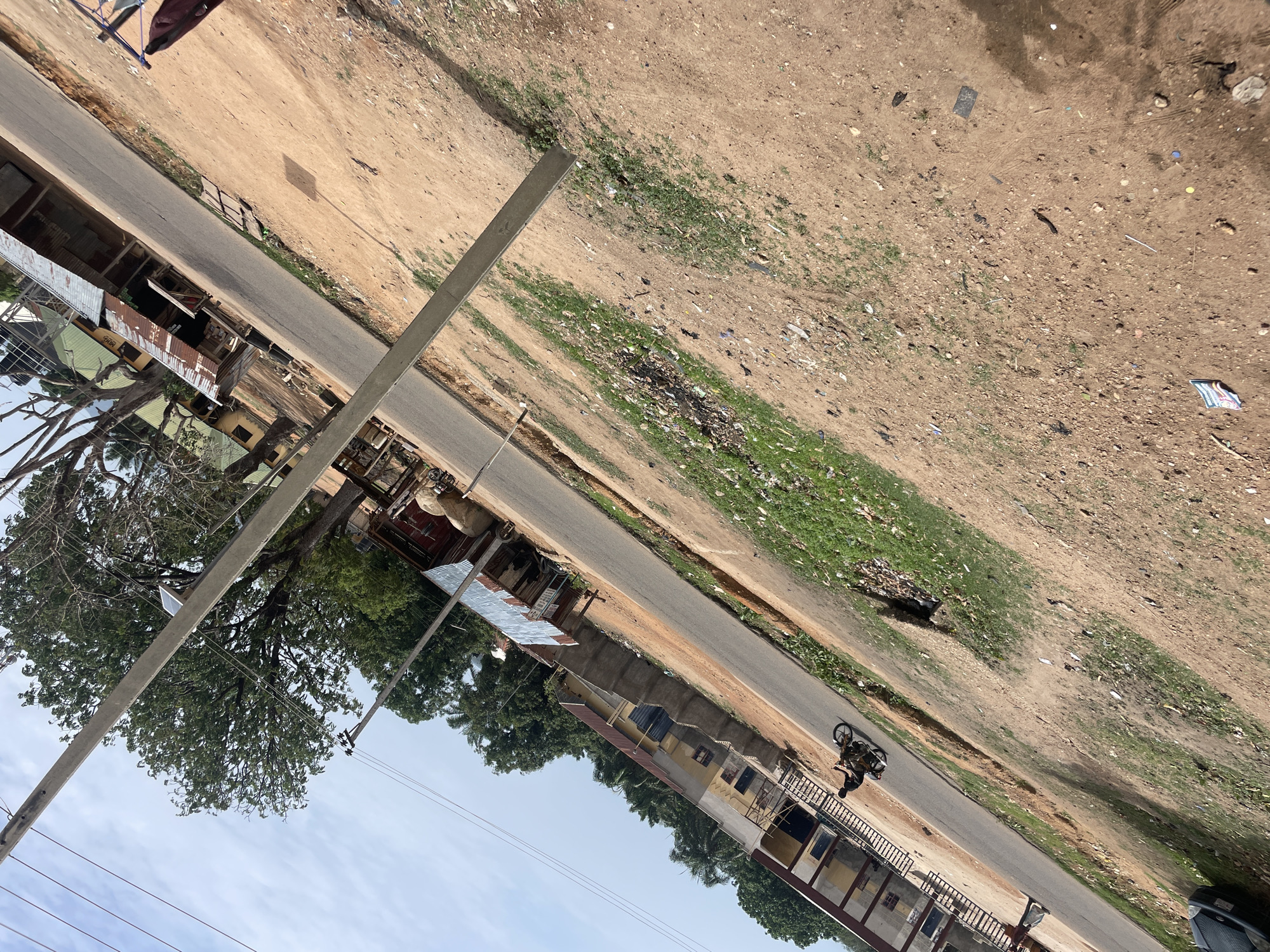 The image depicts a street scene in Mararaba, Nigeria. A paved road runs through the center, flanked by buildings on one side and a dirt area with scattered vegetation and trash on the other. A person is riding a motorcycle on the road. Utility poles are visible along the roadside. The buildings appear to be a mix of residential and commercial structures, some with corrugated metal roofs. The sky is partly cloudy.
