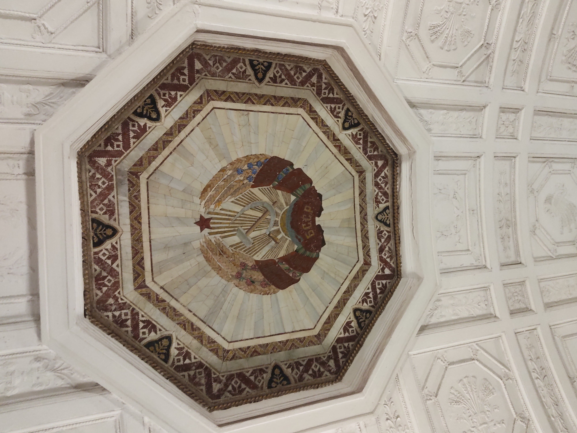 The image shows a ceiling decoration in the form of an octagon, featuring the emblem of the Soviet Union in its center. The emblem consists of a hammer and sickle overlaid on a globe, surrounded by ears of wheat and a red star above. The emblem is made of different colors and material than the background. The background consists of lines radiating from the center, which makes it look like sun rays. The octagon is surrounded by a decorative border with geometric patterns. The ceiling around the octagon is white with relief patterns, suggesting a building of historical or cultural significance, likely a subway station. Given the presence of the Soviet emblem, the image is likely taken in the city of Moscow, Russia, as Moscow's subway stations were often decorated with symbols of the Soviet era. The location could possibly be the Kiyevskaya station, in Moscow, Russia.