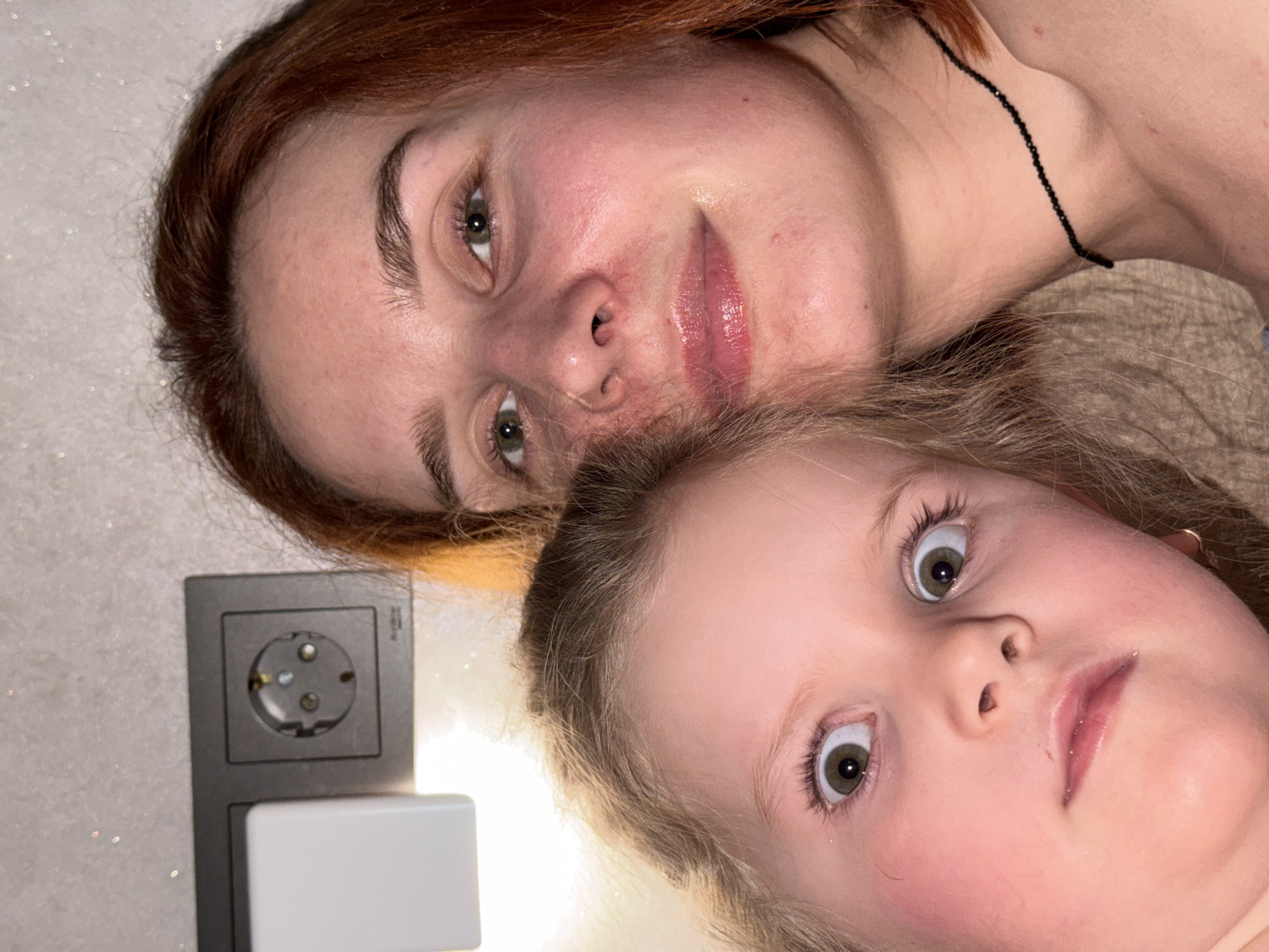 A medium-close shot of two people, likely a mother and daughter, lying down. The woman has reddish-brown hair and a black beaded necklace. The young girl has blonde hair and wide eyes. To the left, there's an electrical outlet with a nightlight. The setting appears to be indoors, possibly a home, but no specific location in Chaikovsky, Russia, can be determined from the image.