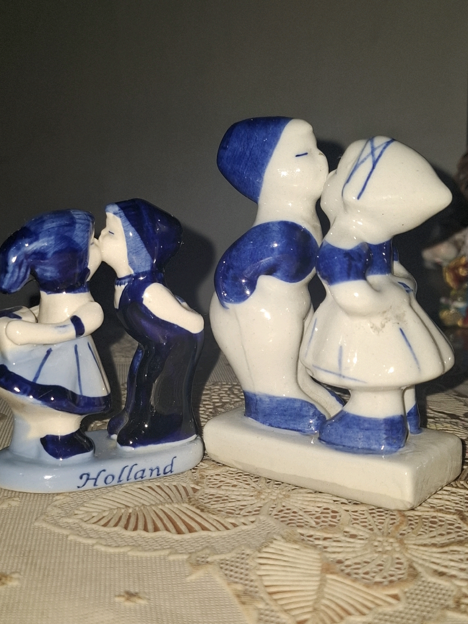 This is a close-up, eye-level shot of two porcelain figurines depicting children kissing, set against a dark, out-of-focus background. The figurines are primarily white with blue detailing, characteristic of Delftware or similar Dutch-style pottery.
The figurine on the left shows a child with dark blue hair tied with a lighter blue band, wearing a light blue dress with darker blue trim. The child is facing left, leaning in to kiss another child.
The figurine on the right appears to be a pair of children also kissing. The child on the left has a blue head covering and is wearing blue and white clothing. The child on the right has a blue head covering and is wearing a white dress with blue lines and trim. Both figures are depicted with closed eyes, suggesting a kiss. The base of this pair of figurines is white.
The two groups of figurines are placed on a light-colored, textured surface that appears to be a lace doily. The word "Holland" is inscribed in blue script on the base of the figurine on the left.
The image does not provide any information about the city of Almaty, Kazakhstan, or any urban environment.