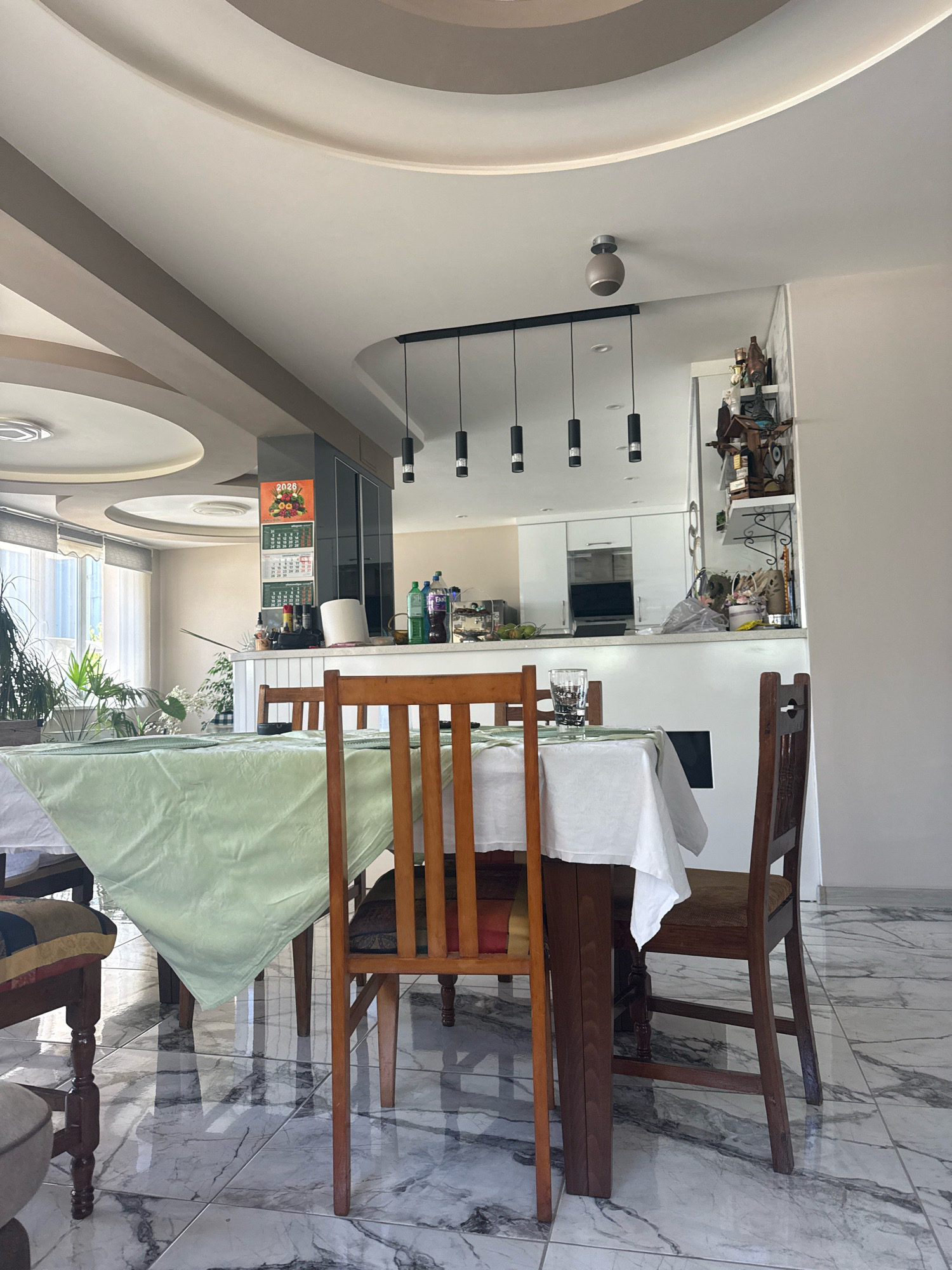 The image depicts an indoor dining area, likely in a residential setting. A rectangular dining table is set with a white tablecloth and a light green runner, covered by a white paper towel dispenser. Two wooden chairs and a wooden bench with upholstered seating are positioned around the table. In the background, a kitchen counter is visible with various items, including bottles and a glass. The room features a modern ceiling design with recessed lighting and a pendant light fixture. The floor is tiled with a marble pattern. The overall impression is of a clean, contemporary living space. No specific location in Varna, Bulgaria is identifiable from the image itself.