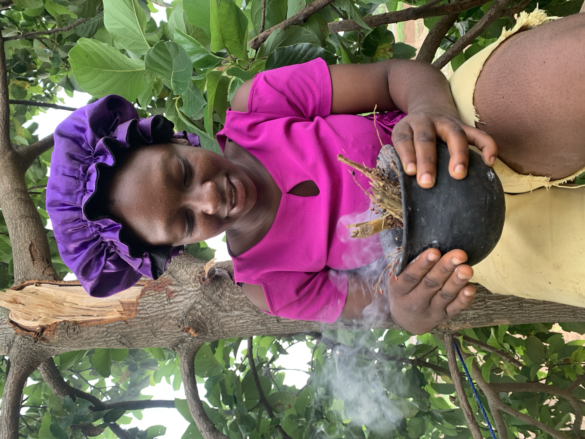 A woman with dark skin, wearing a purple head covering and a pink shirt with a cutout neckline, sits in a tree, holding a black pot containing smoldering material that produces white smoke. The tree has green leaves. The woman is smiling. She wears a yellow skirt or pants. The location is assumed to be Agban, Nigeria.
