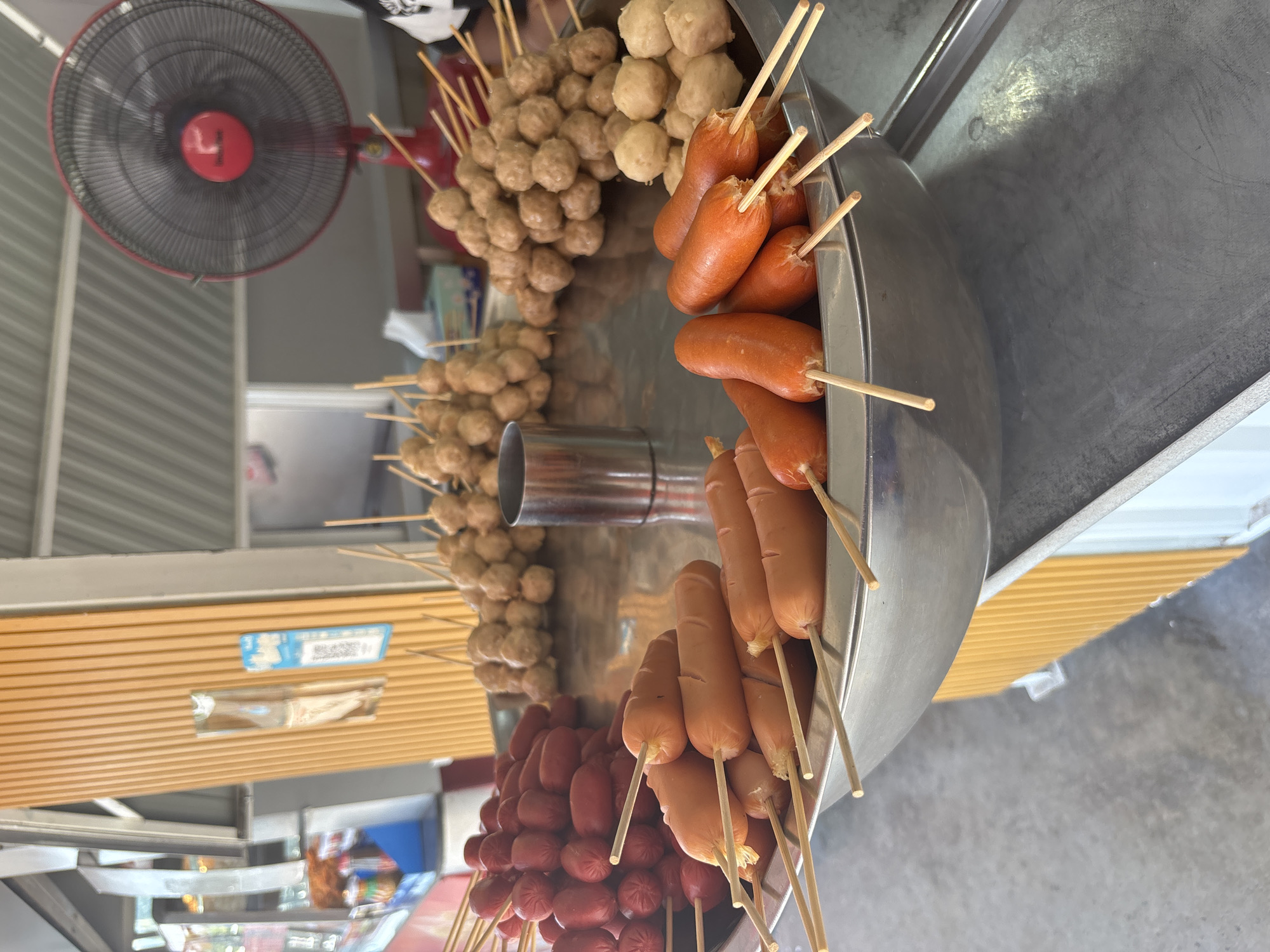 The image shows a street food stall in Baan Bo Phut, Thailand, featuring skewers of sausages and meatballs. The stall is made of stainless steel and is positioned on a grey concrete surface. The skewers are arranged in different sections of the stall: a pile of small red sausages, rows of brown sausages, and clusters of beige meatballs. A red fan is mounted on the side wall, suggesting the warm climate. The wall behind the stall is constructed of horizontal wood slats, and there is a partially visible label.