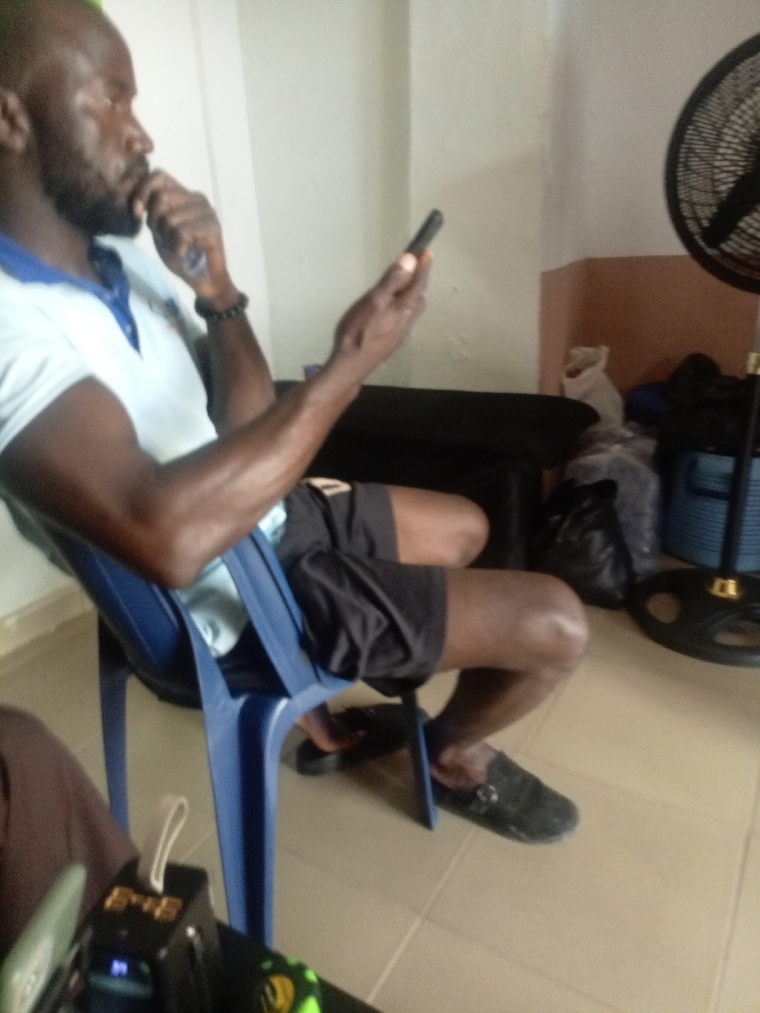 A man with a beard and short hair is seated on a blue plastic chair, looking at his mobile phone. He is wearing a light blue polo shirt and dark shorts. His right hand is raised, with his index finger touching his chin, and his left hand holds the phone. The man is in an indoor setting, likely a room with beige tiled flooring. In the background, a black standing fan is visible, along with a dark-colored piece of furniture, several plastic bags, and what appears to be a shelf with items. The lighting suggests it is daytime, with a light source coming from the left. This scene is located in the city of Jos, Nigeria.