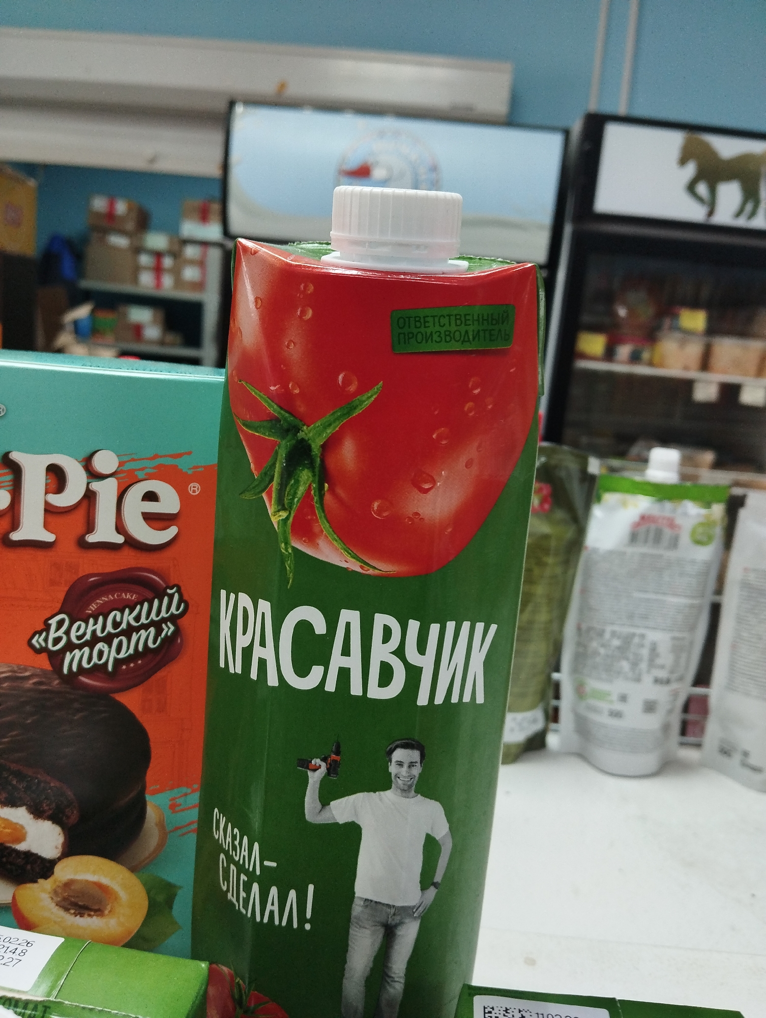 The image captures a retail setting in Krasnogorskoye, Russia, where a carton of "Krasavchik" tomato juice is prominently displayed alongside a box of "Pie" brand Viennese cake. The juice carton features a large image of a tomato and a man holding a drill. In the background, shelving with various grocery items is visible, along with a refrigerated display case showcasing different products. The scene suggests a typical grocery store interior.