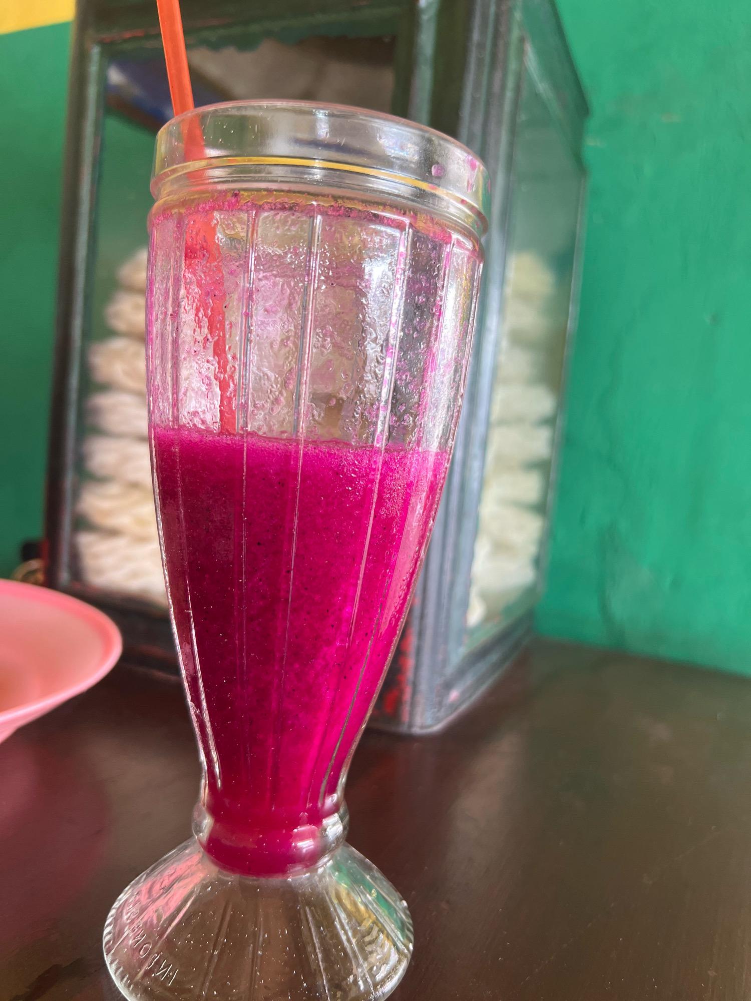 An extreme close-up shot focuses on a tall, fluted glass filled with a vibrant pink, frothy beverage, presumably dragon fruit juice, and a red straw. The glass sits on a dark, reflective surface, likely a table. To the left, a portion of a pink plate is visible. In the background, a transparent display case contains stacked white food items, and a bright green wall serves as the backdrop. The scene is set in a casual establishment in Sayegan, Indonesia.