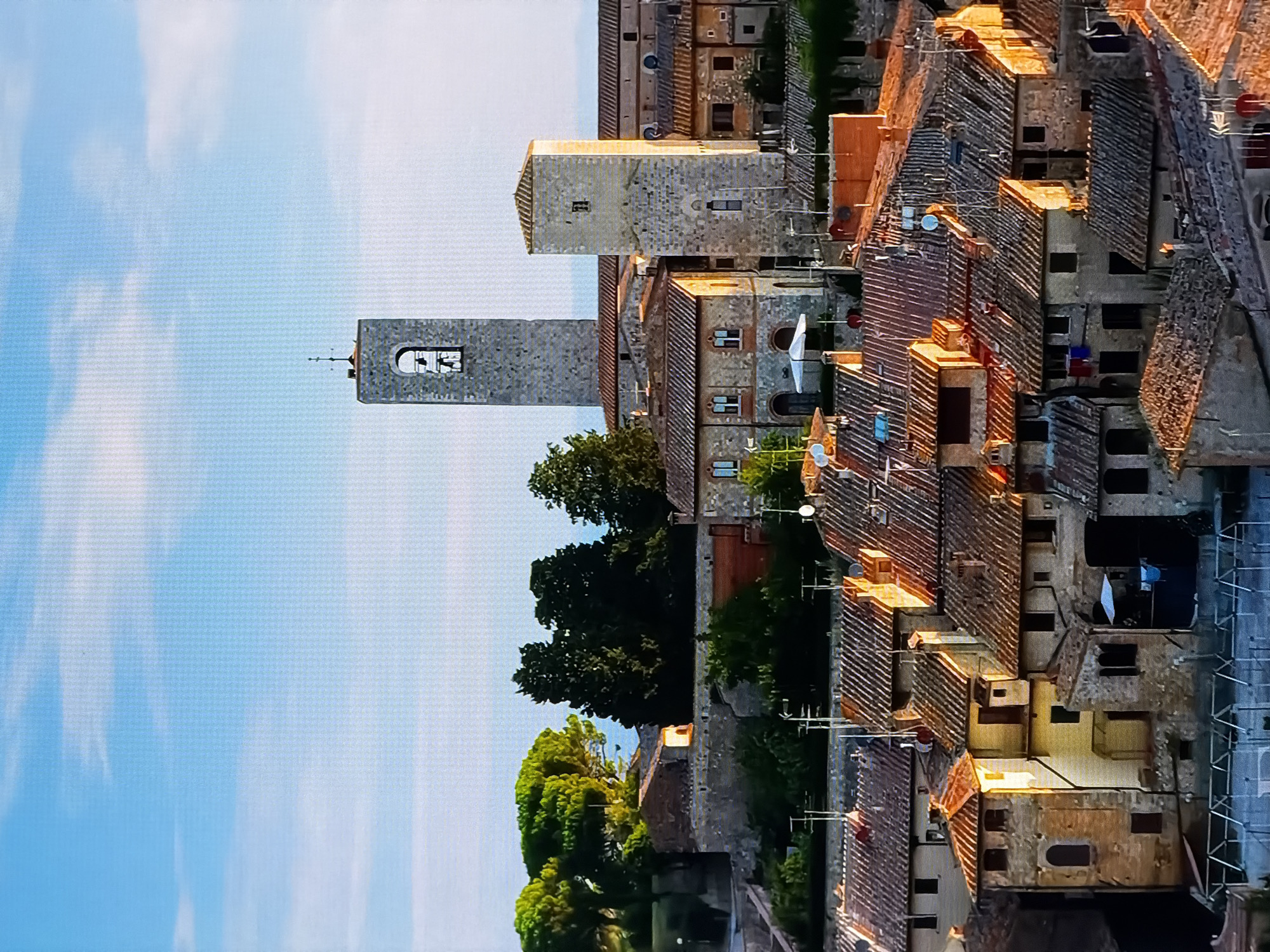 This is a high-angle, full-shot photograph of a European town, likely in Tuscany, Italy. The image features a cluster of buildings with terracotta roofs, varying in height and architectural style. Dominating the skyline are two stone towers; a taller, narrower tower with a bell visible in its arched opening, and a shorter, squarer tower. The buildings are constructed from stone and brick, with visible windows and doorways. Some buildings have laundry hanging out, suggesting residential use. Trees, some lush and green, are interspersed with the buildings, and the scene is set against a bright blue sky with scattered white clouds. The lighting suggests it is either early morning or late afternoon, with the sun casting warm light on some of the structures. There is no indication that this scene is located in Chirchik, Uzbekistan.