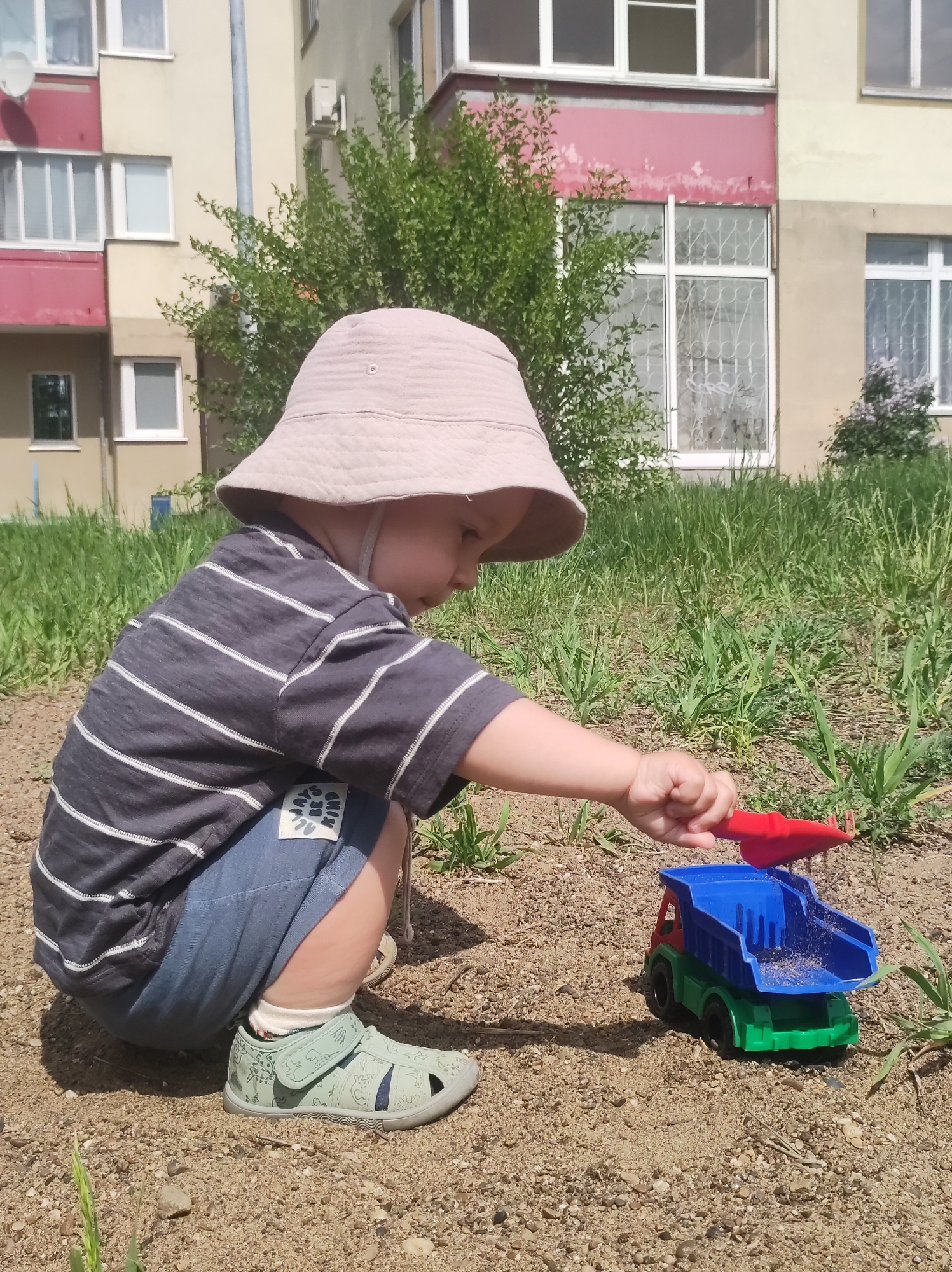 A child is playing in the sand with a toy truck and a red shovel. The child, wearing a beige bucket hat, a striped gray shirt, blue shorts, and green sandals, is squatting in the sand and holding the shovel over the truck's blue bed. The toy truck is green, blue, and red. The sand is mixed with small rocks and sparse vegetation. Behind the child and the sand are tall blades of grass and a building with windows and a bush. Based on the available information, the location is the city of Chaikovsky, Russia.