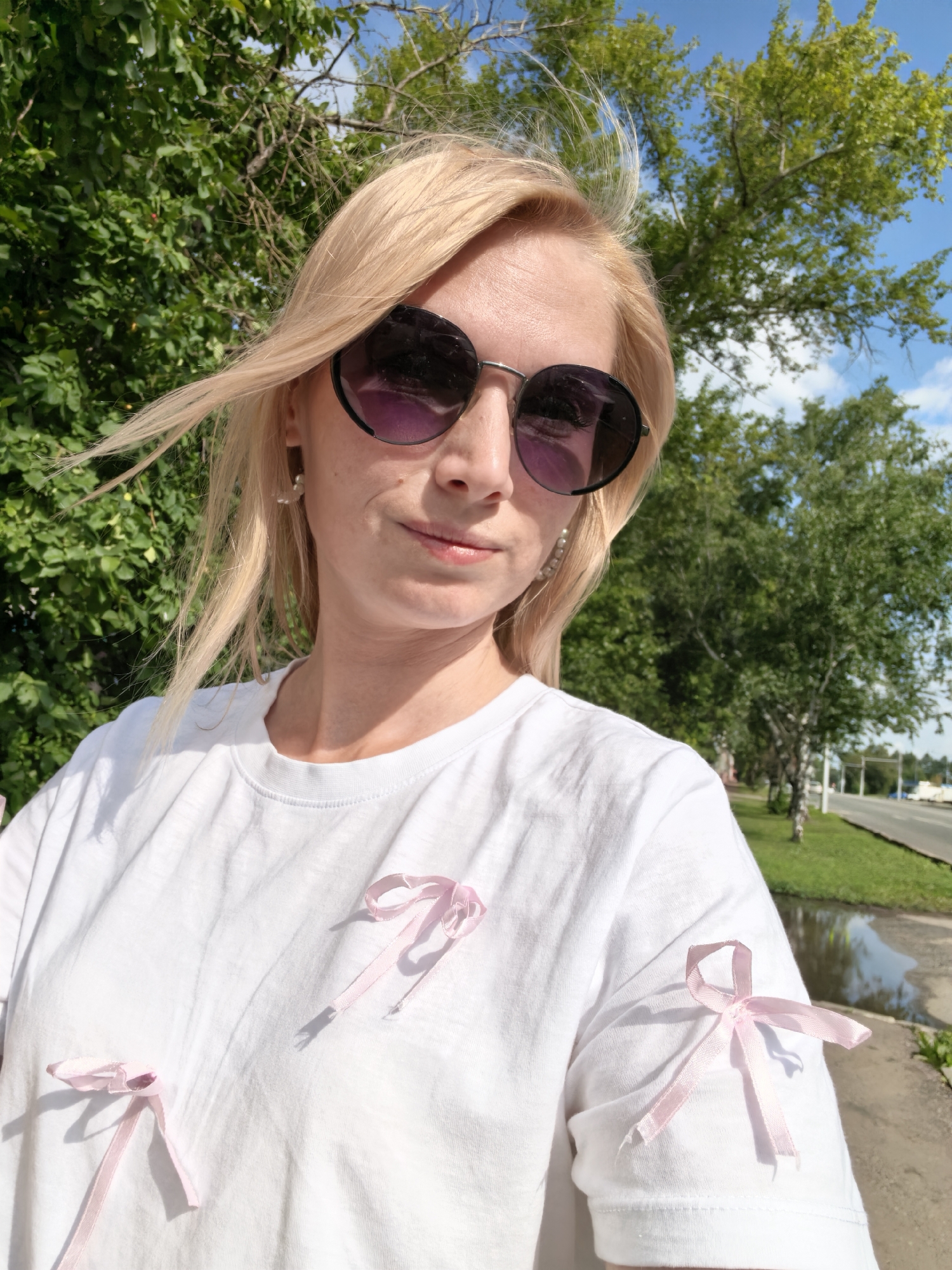 A Caucasian woman with blonde hair and sunglasses stands outdoors on a sunny day, with trees and a road visible in the background. She is wearing a white t-shirt adorned with several small pink bows. The setting appears to be a roadside area in Sterlitamak, Russia, suggested by the presence of a paved road, sidewalk, grassy verge, and foliage consistent with the region's vegetation. The lighting indicates it is daytime, and the sky is blue with some clouds.