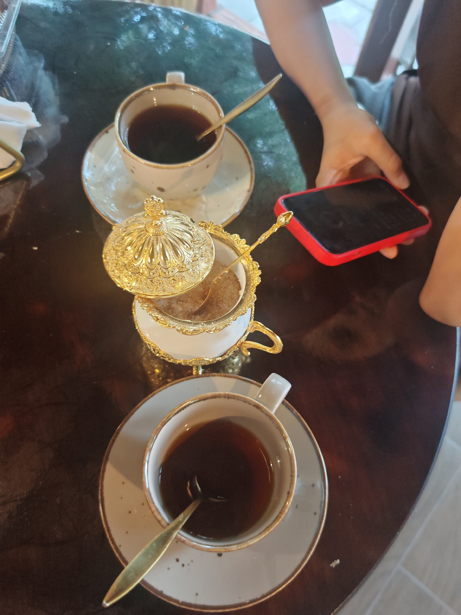 A high-angle shot captures a dark wooden table with two cups of coffee and a sugar bowl. In the foreground, a cup and saucer hold dark coffee with a gold spoon resting inside. To the left, a decorative white and gold sugar bowl is filled with light brown sugar, also with a gold spoon. Behind the sugar bowl, another cup and saucer with coffee and a spoon are visible. A person's arm and hand are in the upper right corner, holding a red-cased smartphone. The scene appears to be indoors, likely a cafe, with a glimpse of what might be tile flooring in the background. The lighting suggests it could be daytime, possibly late afternoon. No visible text is present in the image. The overall mood is relaxed, perhaps a break for coffee and conversation or a moment of personal reflection.