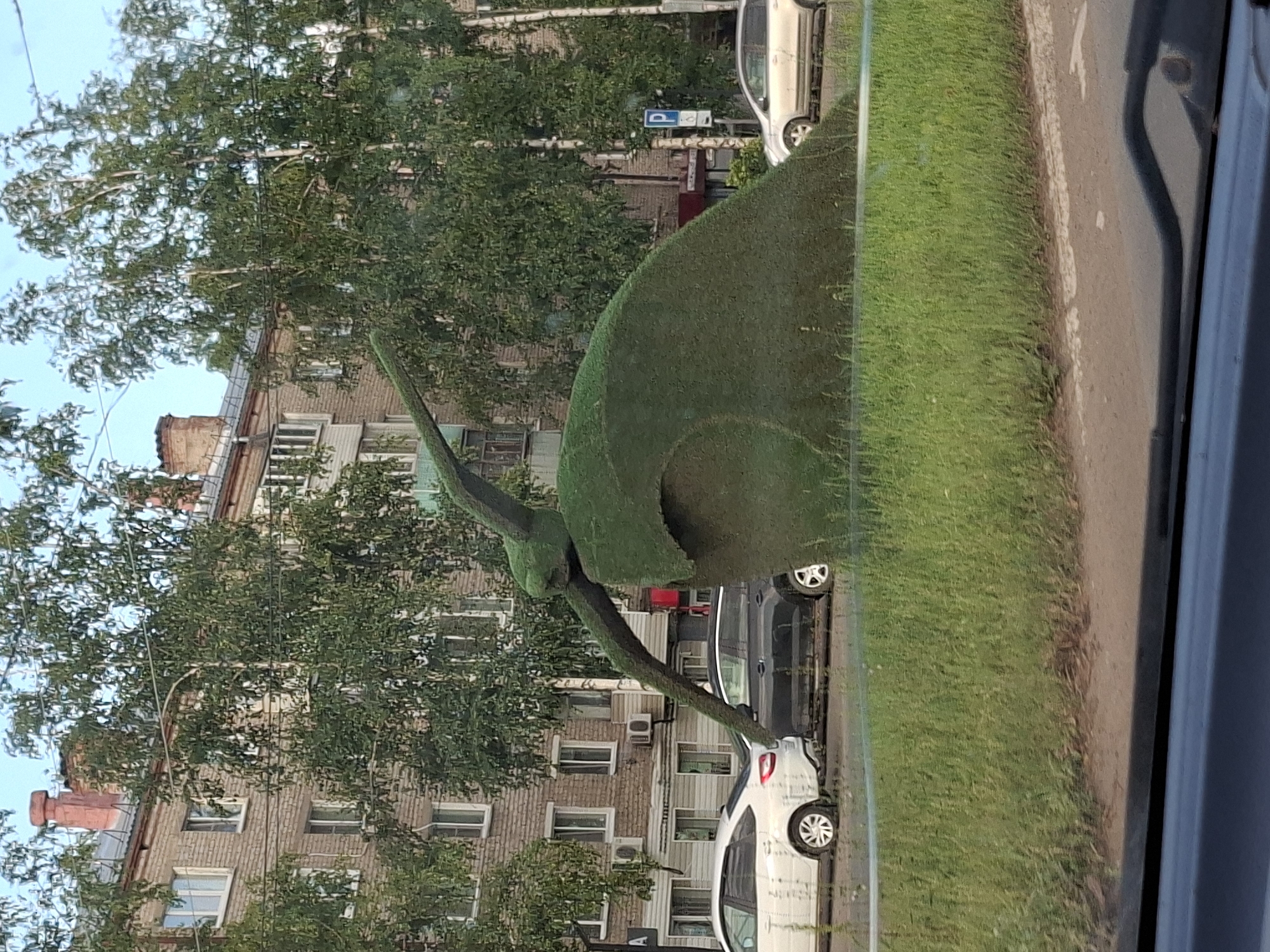 This image, captured in Chaikovsky, Russia, shows a topiary sculpture resembling a snail. The sculpture is green and is situated on a grassy area next to a road. In the background, there is a multi-story brick building with many windows and balconies, suggesting an urban or suburban residential area. Trees with green foliage are visible in front of the building and behind the sculpture. Two cars are parked on the side of the road; one is dark and the other is white. The time of day appears to be daytime, with overcast skies. The image is taken from a low angle, possibly from within a vehicle, looking out. The orientation of the image is sideways. No text is clearly visible in the image.