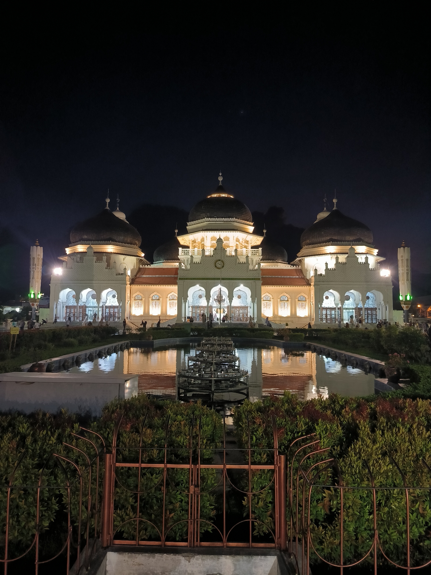 The image depicts the Baiturrahman Grand Mosque in Banda Aceh, Indonesia, at night. The mosque is illuminated by warm interior lights, highlighting its white facade, multiple domes, and minarets. A reflective pool of water in front of the mosque mirrors the building and its lights, creating a symmetrical view. The foreground features a dark metal fence and lush green bushes. Several people are visible in the distance, walking around the mosque grounds. The sky above is dark and clear.