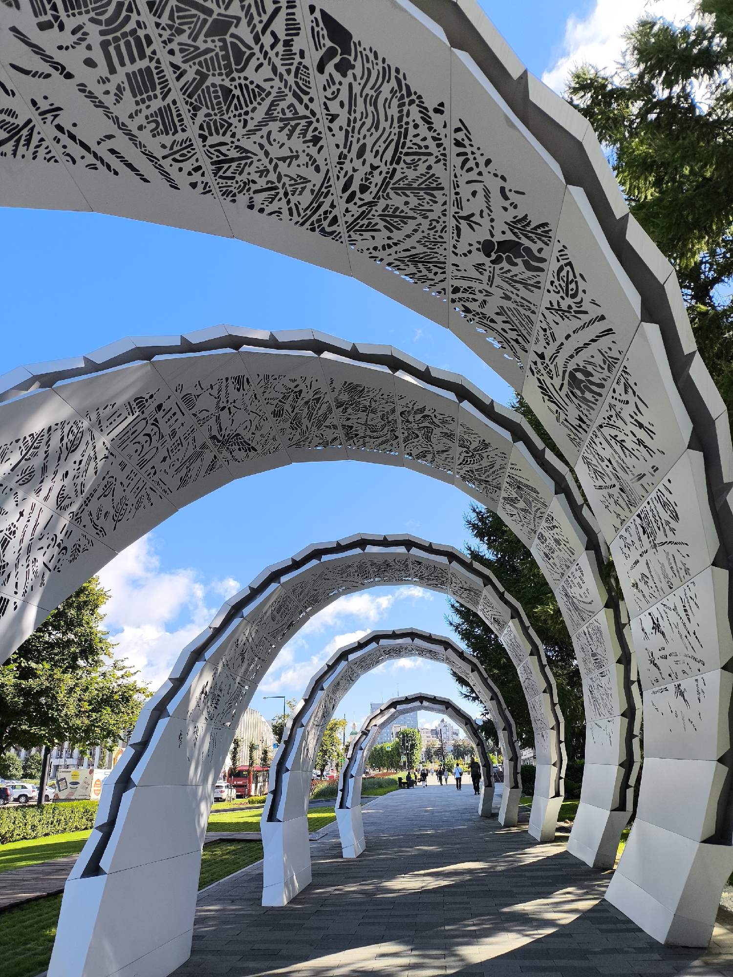 This is a medium-shot, eye-level, outdoor photograph of a walkway under a series of white, modern, arched sculptures in Perm, Russia. The sculptures are composed of geometric segments with intricate, cutout patterns on their upper surfaces, allowing sunlight to filter through. The walkway itself is paved with gray, rectangular stones.
To the left of the walkway, there is a neatly manicured lawn, a wooden deck, and trees. Further beyond the lawn, a street is visible with cars, buildings, and people walking. The sky is bright blue with scattered white clouds. To the right of the walkway, trees with green foliage are present.
In the background, along the walkway, several people are walking away from the camera, appearing as small figures in the distance. The architecture suggests a public space, possibly a park or pedestrian zone in a city. The overall atmosphere is bright and sunny. The sculpture is located on the embankment of the Kama River in Perm.