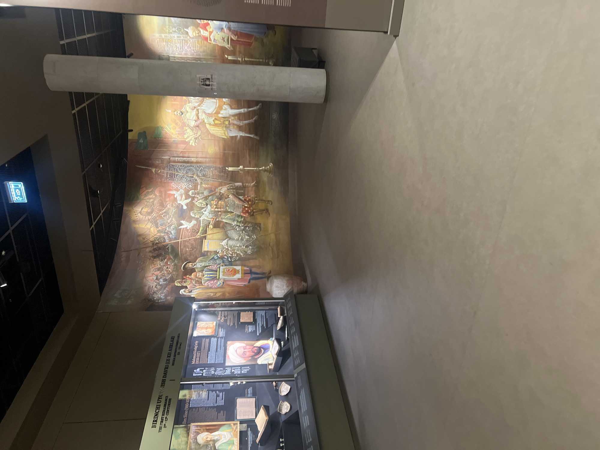 The image shows an indoor exhibit, possibly a museum, with a large mural on the back wall depicting a historical scene with soldiers, horses, and banners. In the foreground, there is a display case with artifacts and text panels. The floor is a light-colored concrete. A large, fluted concrete column stands to the right of the mural. The lighting appears to be artificial. The text on the display case is in an unfamiliar language. The overall impression is that of a historical or cultural exhibition.