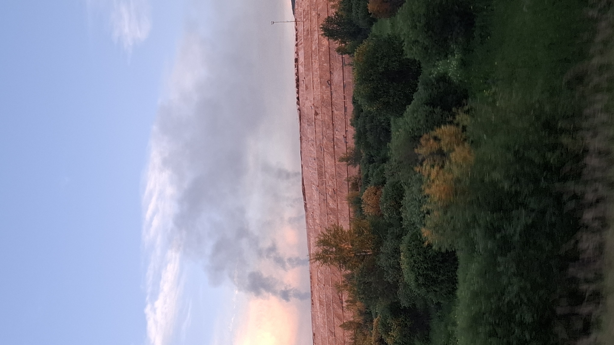 This media file depicts a landscape scene, likely taken from a moving vehicle due to the motion blur. The image is oriented vertically, with the sky occupying the left half and the ground the right. The sky is a pale blue with scattered, wispy white clouds that are tinged with pink and orange hues, suggesting it is either sunrise or sunset.
The right side of the image shows a densely wooded area with various shades of green foliage, interspersed with a few trees displaying autumn colors of yellow and orange. The ground appears to be a mix of grass and possibly some undergrowth. In the middle ground, a long, flat structure covered in a textured, reddish-pink material is visible. This structure runs horizontally across the frame and appears to be a man-made object. A single lamppost stands at the far end of this structure.
The overall mood is serene, with the soft colors of the sky and the natural elements of the trees. There is no visible text in the image. The setting is likely rural or semi-rural, given the open space and presence of trees. The time of day is either dawn or dusk.