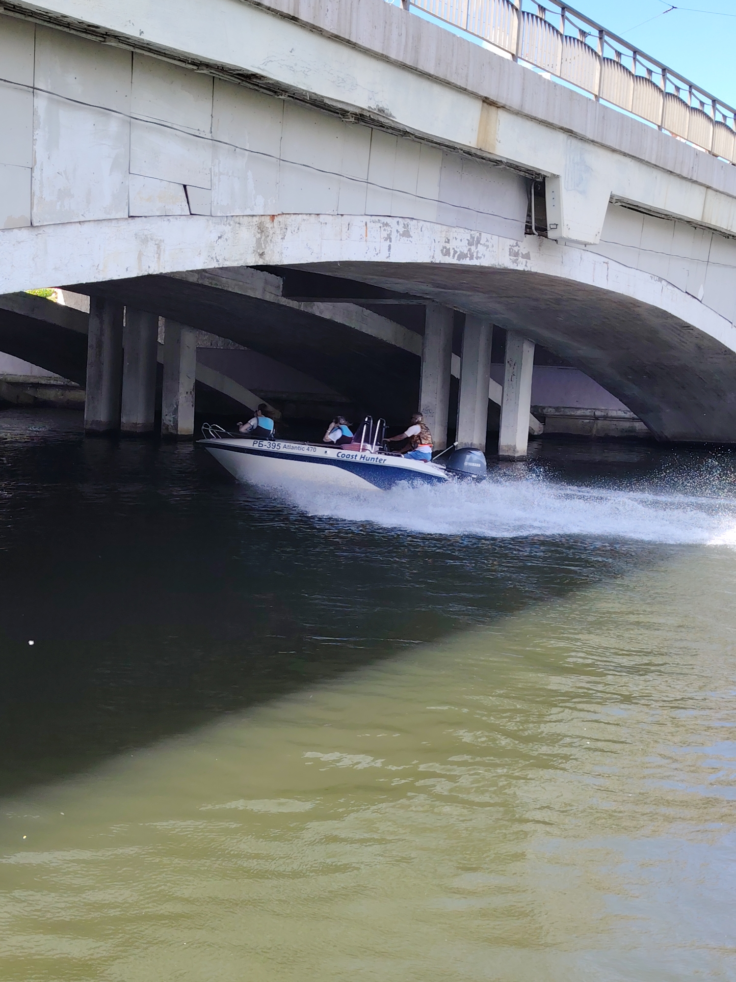 This image depicts a motorboat navigating a waterway under a concrete bridge in Minsk, Belarus, during what appears to be a clear day.
The primary subjects are a white and blue motorboat, three individuals aboard it, and the large concrete bridge spanning the water. The boat, identified by the text "РБ-395 Atlantic 470" and "Coast Hunter" on its hull, is powered by a Yamaha outboard engine, which is clearly visible at the stern. It is actively moving, leaving a distinct white wake of disturbed water behind it.
Of the three people on the boat, one is at the helm, operating the vessel, while the other two are seated as passengers. All three appear to be wearing life vests.
The bridge itself is a substantial concrete structure featuring multiple arched sections and sturdy support pillars rooted in the water. The underside of the bridge shows signs of wear and tear; several paneling sections appear loose, cracked, or missing, exposing the inner structure and some wiring. Metal railings are visible along the top of the bridge, suggesting it carries a road or pathway.
The water beneath the bridge displays varying shades, from very dark in the deep shadows directly under the structure to lighter, greenish-brown tones where it catches more light. The clear daylight is evident from the bright reflections on the water and the defined shadows cast by the bridge. There are no strong indications of specific emotions from the people, and the weather seems calm and sunny.