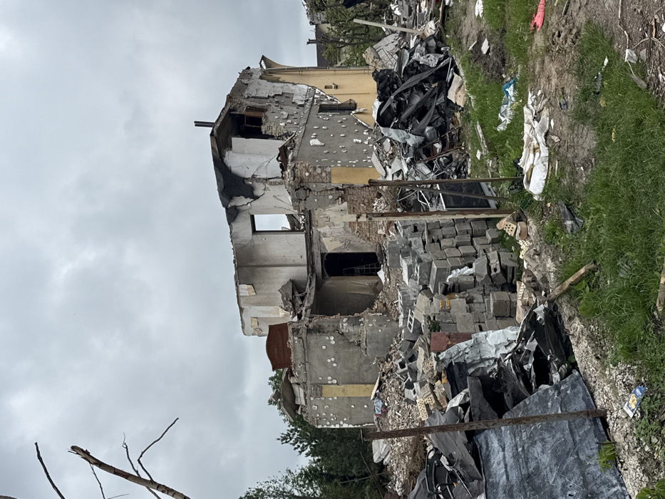 A severely damaged two-story building stands amidst debris. The structure is heavily scarred with holes and missing sections, revealing inner layers and collapsed portions. Rubble, including brick piles and twisted metal, surrounds the building. Vegetation flanks the foreground, contrasting with the destruction. The sky is overcast, contributing to the somber atmosphere.