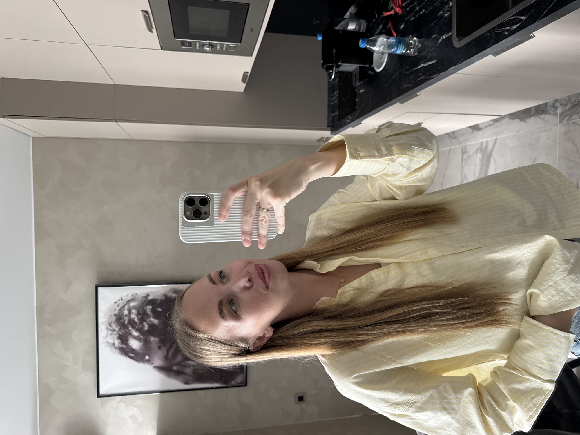 A young woman with long, straight blonde hair, wearing a light yellow striped long-sleeved shirt, takes a mirror selfie in what appears to be a modern kitchen and living space. She holds a white smartphone with a ridged case in her left hand, which has a ring on the ring finger. Her head is slightly tilted, and she has a calm, neutral expression.
The background features a contemporary kitchen area with light-colored upper and lower cabinets and a built-in dark microwave oven. A black marble-patterned countertop holds a coffee machine and a plastic water bottle. The walls are light beige with a textured finish. To the left of the woman, a framed black and white piece of art depicting a textured, possibly abstract, subject hangs on the wall. The flooring is a light-colored tile, possibly marble or ceramic. The setting suggests an indoor environment, likely an apartment or hotel room in Sochi, Russia. The lighting is soft and even, indicating daytime.