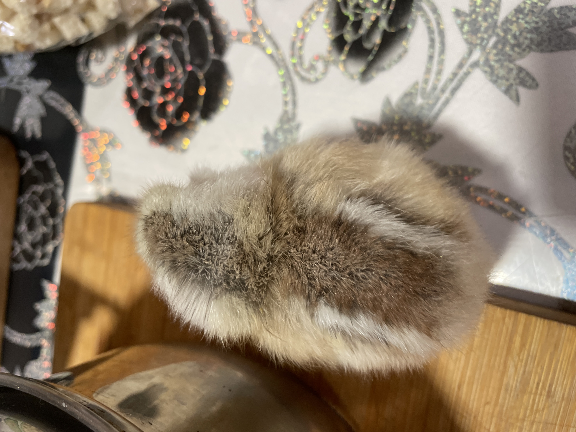 A close-up shot shows a furry, oval object, possibly a pom-pom or a small animal, resting on a wooden surface next to a metallic object. The background consists of a patterned surface, possibly a tablecloth with floral designs in black, silver, and white. There is no evidence to support a location in the city of Zlatograd, Bulgaria. No people, actions, or interactions are visible.